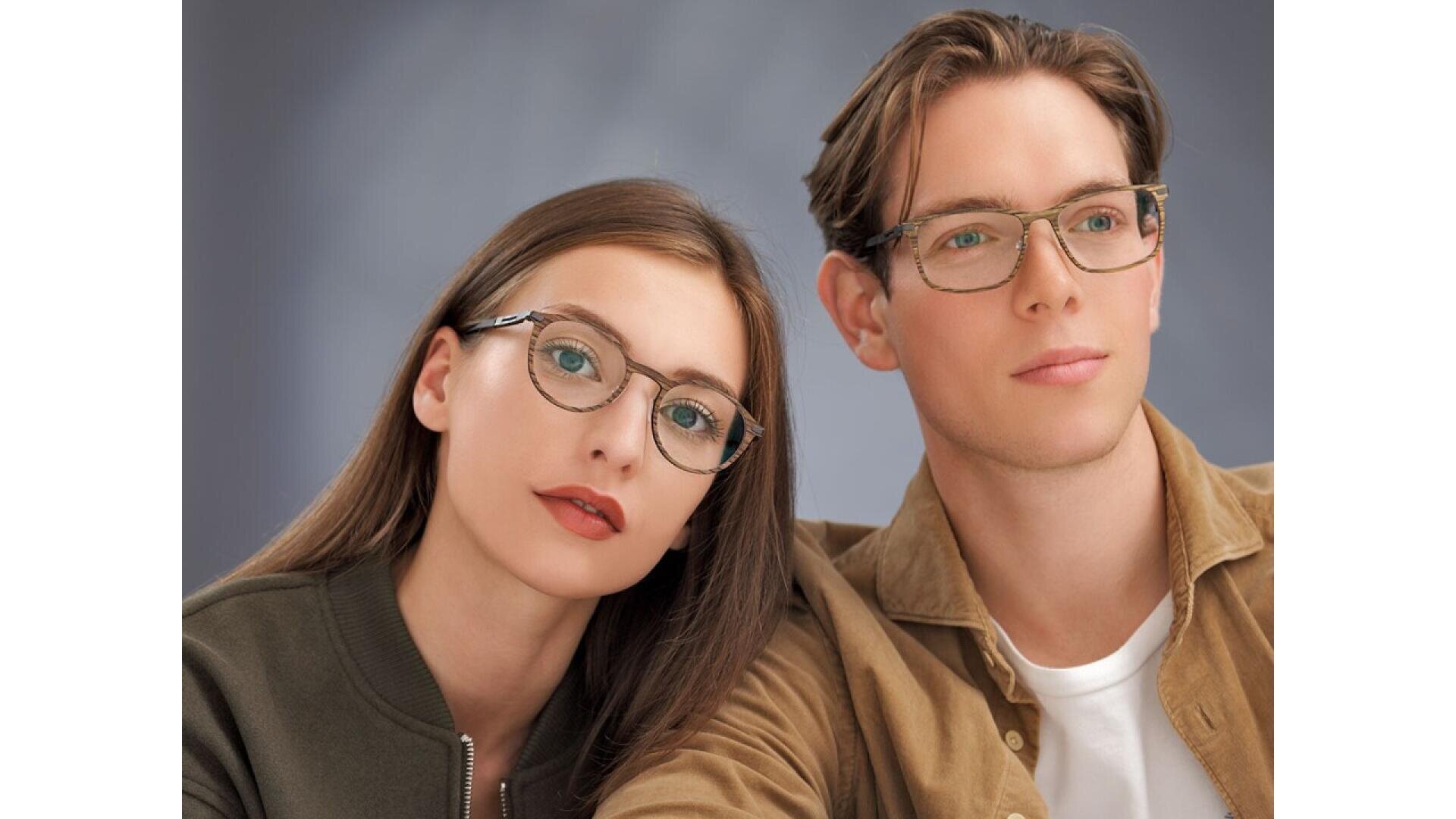 A young woman and man with light skin, both wearing glasses, sit closely together and look slightly off-camera. The woman has long brown hair and wears a green jacket; the man has short hair and wears a tan jacket over a white shirt.