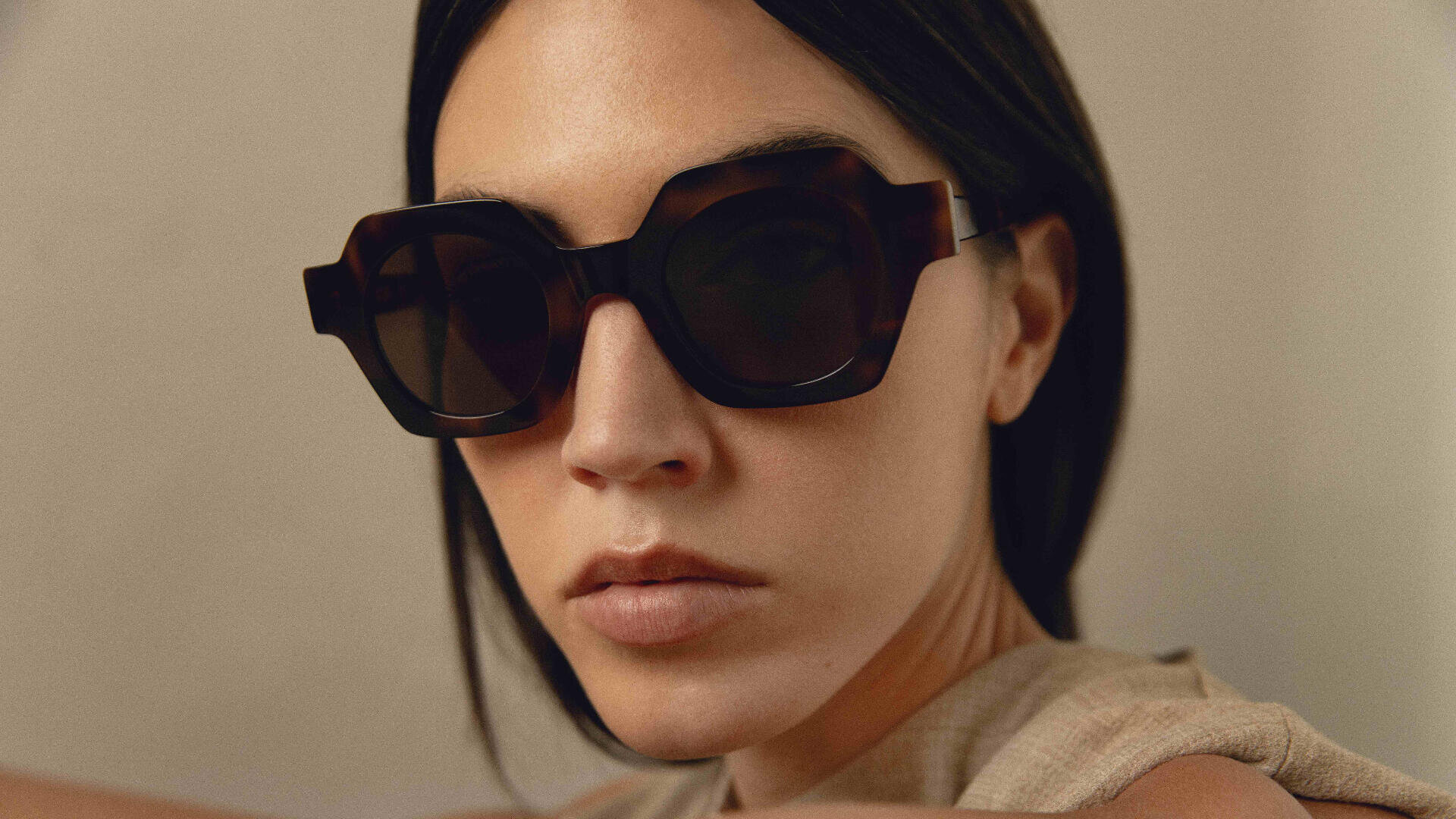 A woman with straight dark hair wears large, geometric, dark sunglasses and a sleeveless beige top, looking directly at the camera with a neutral expression against a plain background.