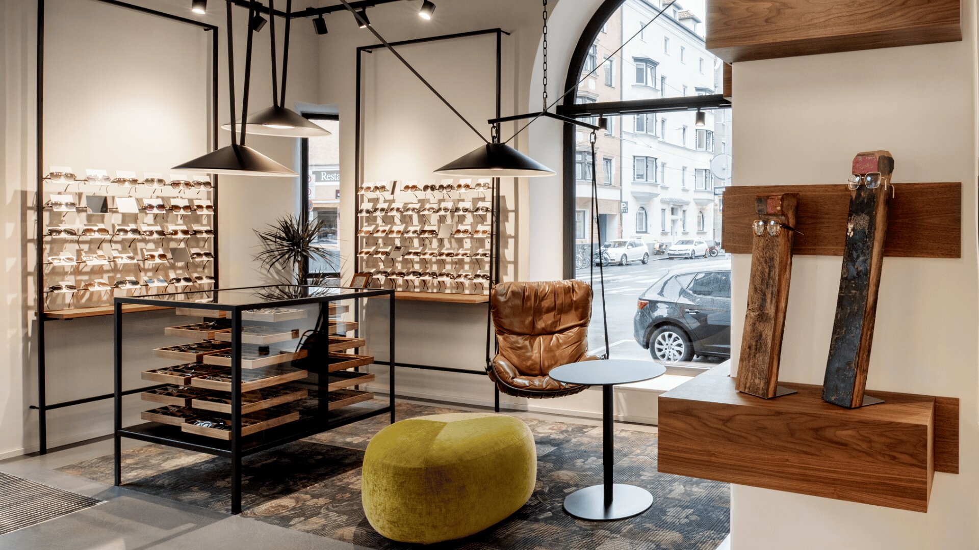 Ein modernes Brillengeschäft mit Brillen, die in Holzregalen ausgestellt sind, einem braunen Ledersessel, einem runden Tisch, einem grünen Hocker und großen Fenstern mit Blick auf geparkte Autos und Gebäude draußen.