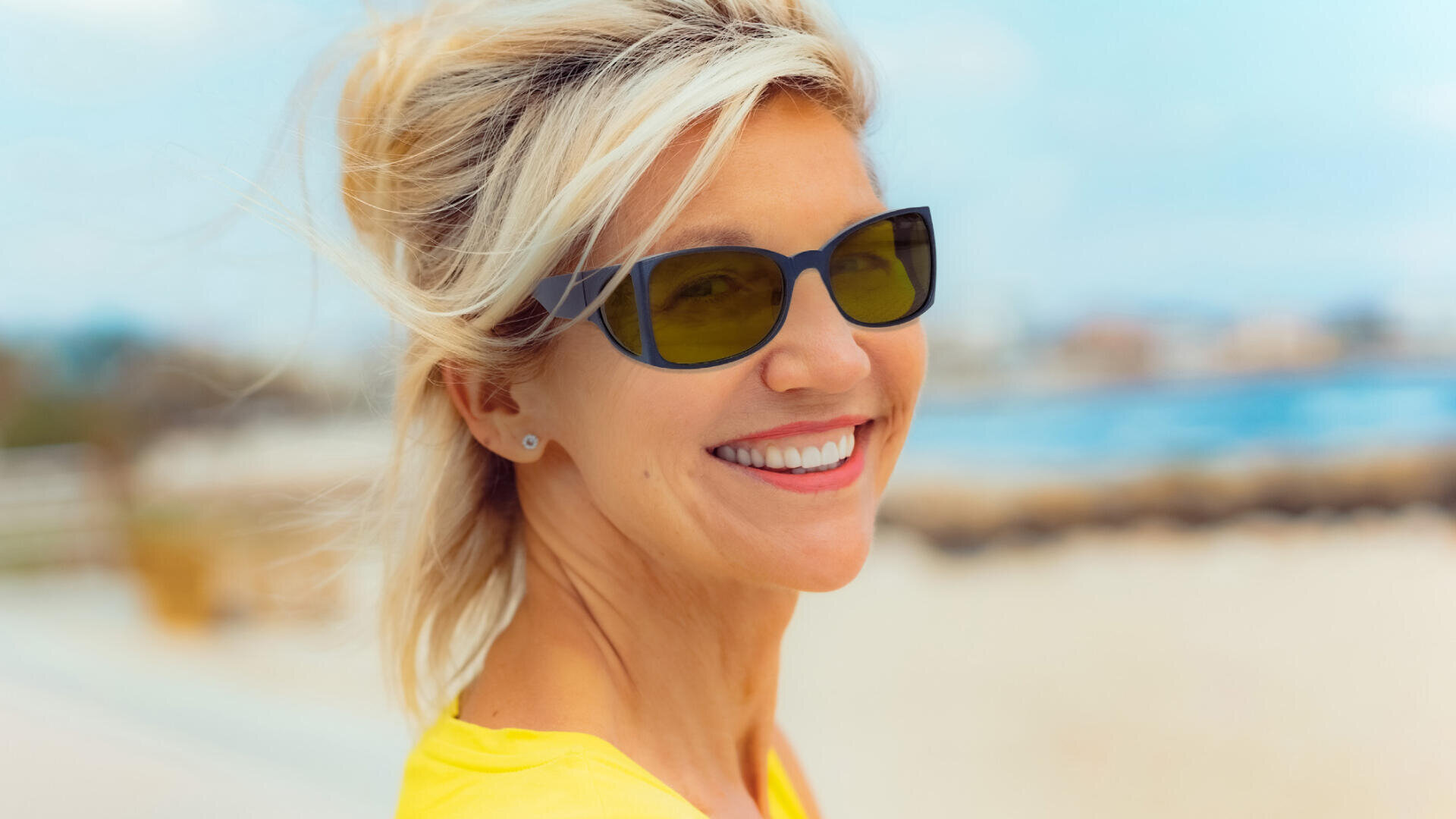 Eine lächelnde Frau mit blondem Haar, einer schwarzen Sonnenbrille und einem gelben Oberteil steht an einem sonnigen Tag im Freien, im Hintergrund sind ein verschwommener Strand und das Meer zu sehen.