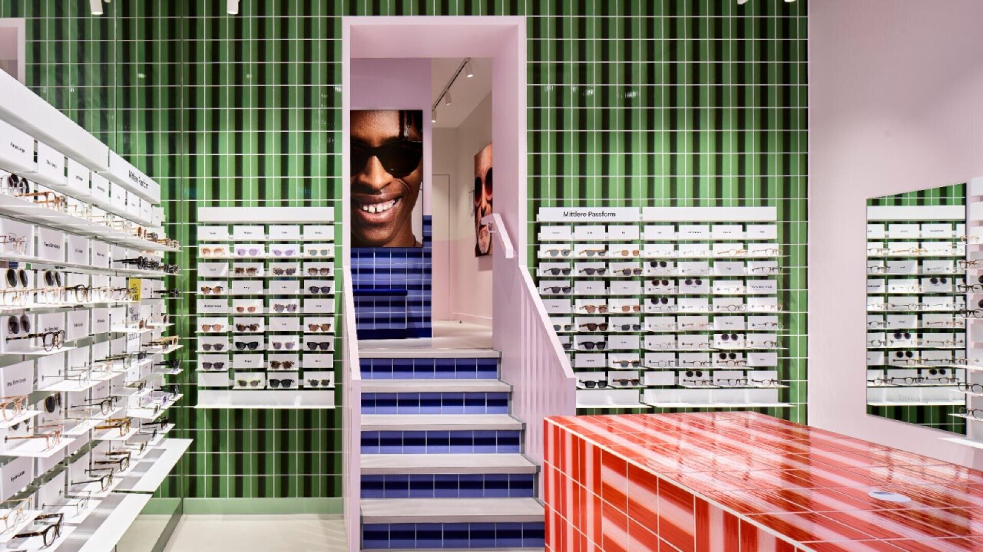 A modern eyewear store features green and white tiled walls, rows of glasses displayed on shelves, a red and white striped counter, and blue-tiled stairs leading to a large photo of a smiling person wearing sunglasses.