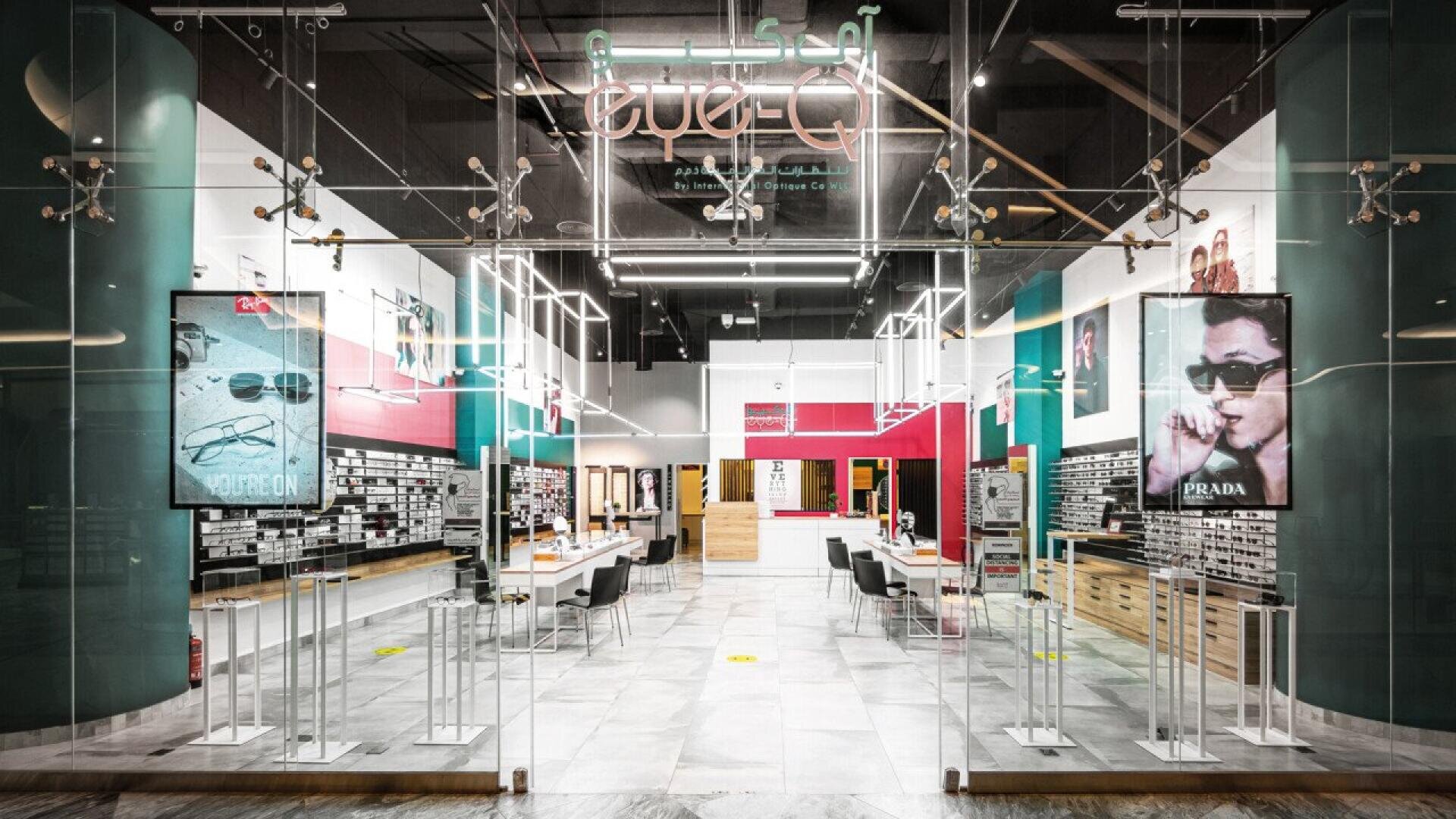 A modern eyewear store with glass doors, bright lighting, display shelves filled with eyeglasses, and large posters featuring models wearing sunglasses on each side of the entrance.