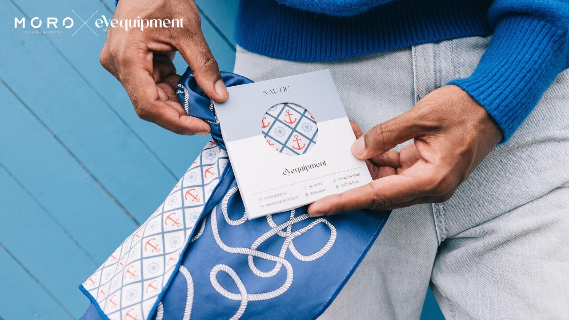 A person in a blue sweater and light jeans holds a blue patterned scarf and a product card with nautical designs featuring red and blue anchors and ship wheels. Logos for MGRO and eyequipment appear in the top left corner.