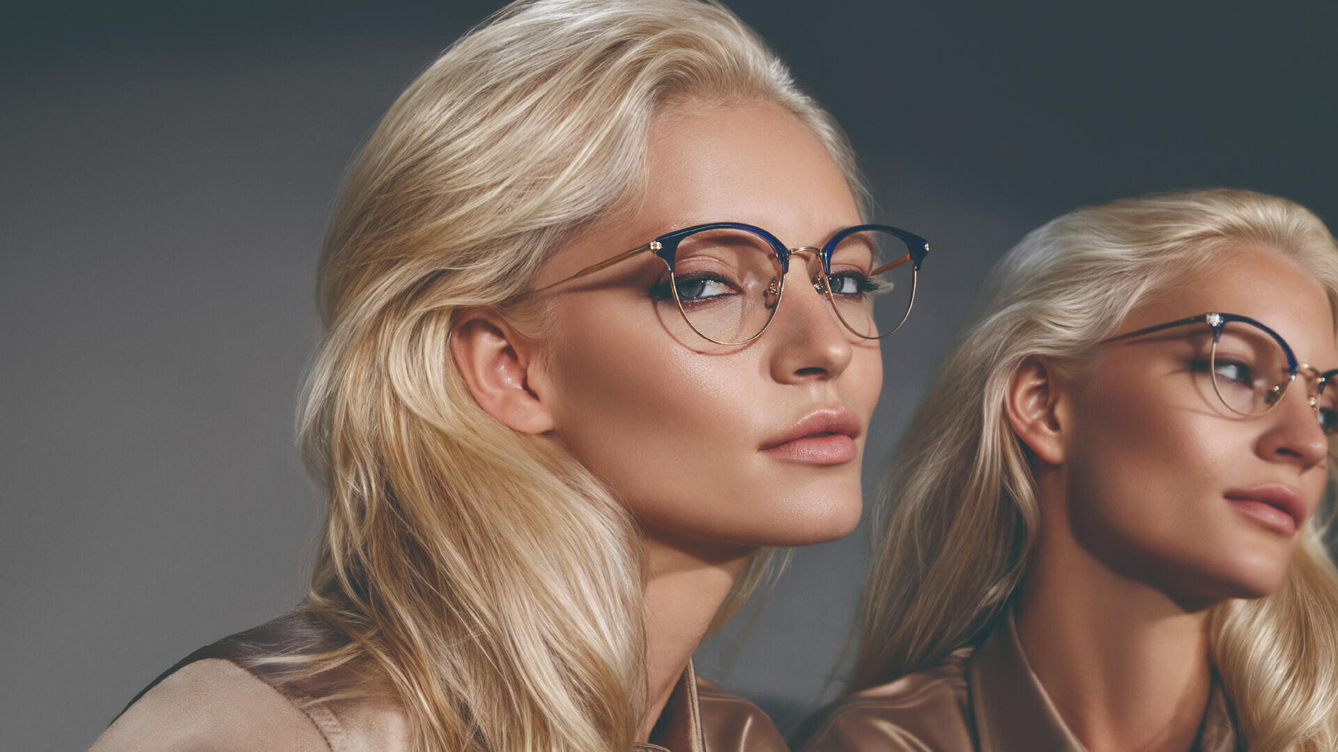 Eine Frau mit langen, blonden Haaren und Brille blickt selbstbewusst in die Kamera. Sie trägt ein seidiges, braunes Hemd. Ihr Spiegelbild neben ihr unterstreicht ihr stilvolles Auftreten.