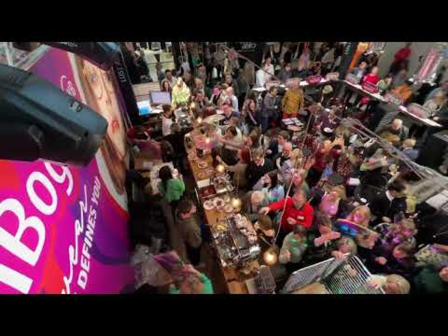 A crowded market hall with many people clustered around food stalls, seen from above. Bright signs and various dishes can be seen as people interact and shop.