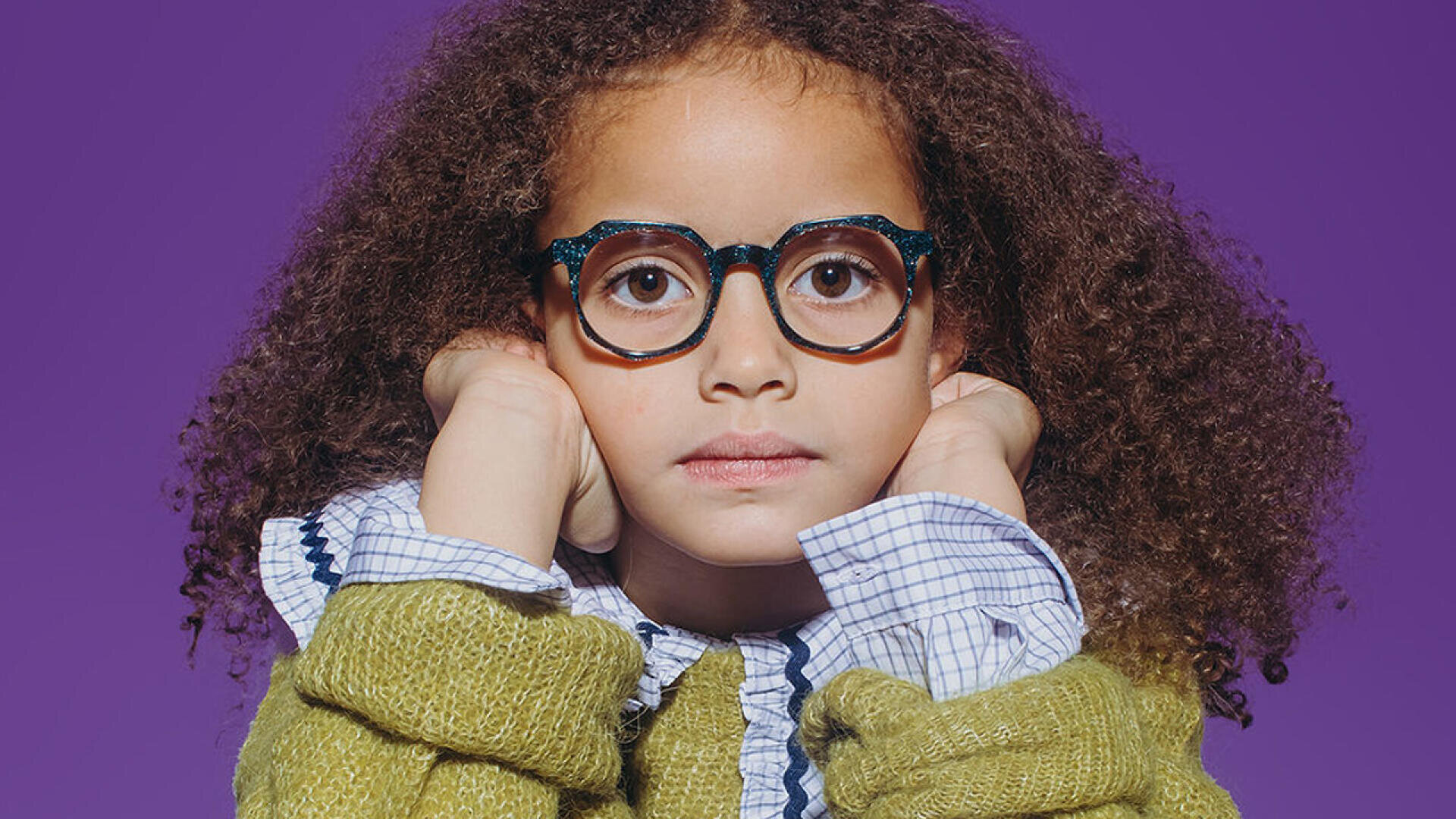 Ein junges Mädchen mit lockigem Haar, einer runden Brille und einem grünen Pullover sitzt mit den Händen auf den Wangen da und blickt vor einem violetten Hintergrund direkt in die Kamera.