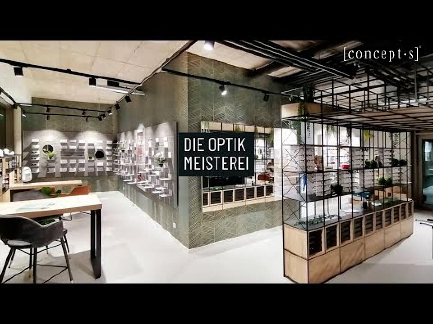Modernes Interieur eines Optikergeschäfts mit stilvollem Dekor, Brillen in Regalen und einem Schild mit der Aufschrift "DIE OPTIK MEISTEREI" in der Mitte. Das Geschäft hat ein offenes, minimalistisches Design und industrielle Elemente.