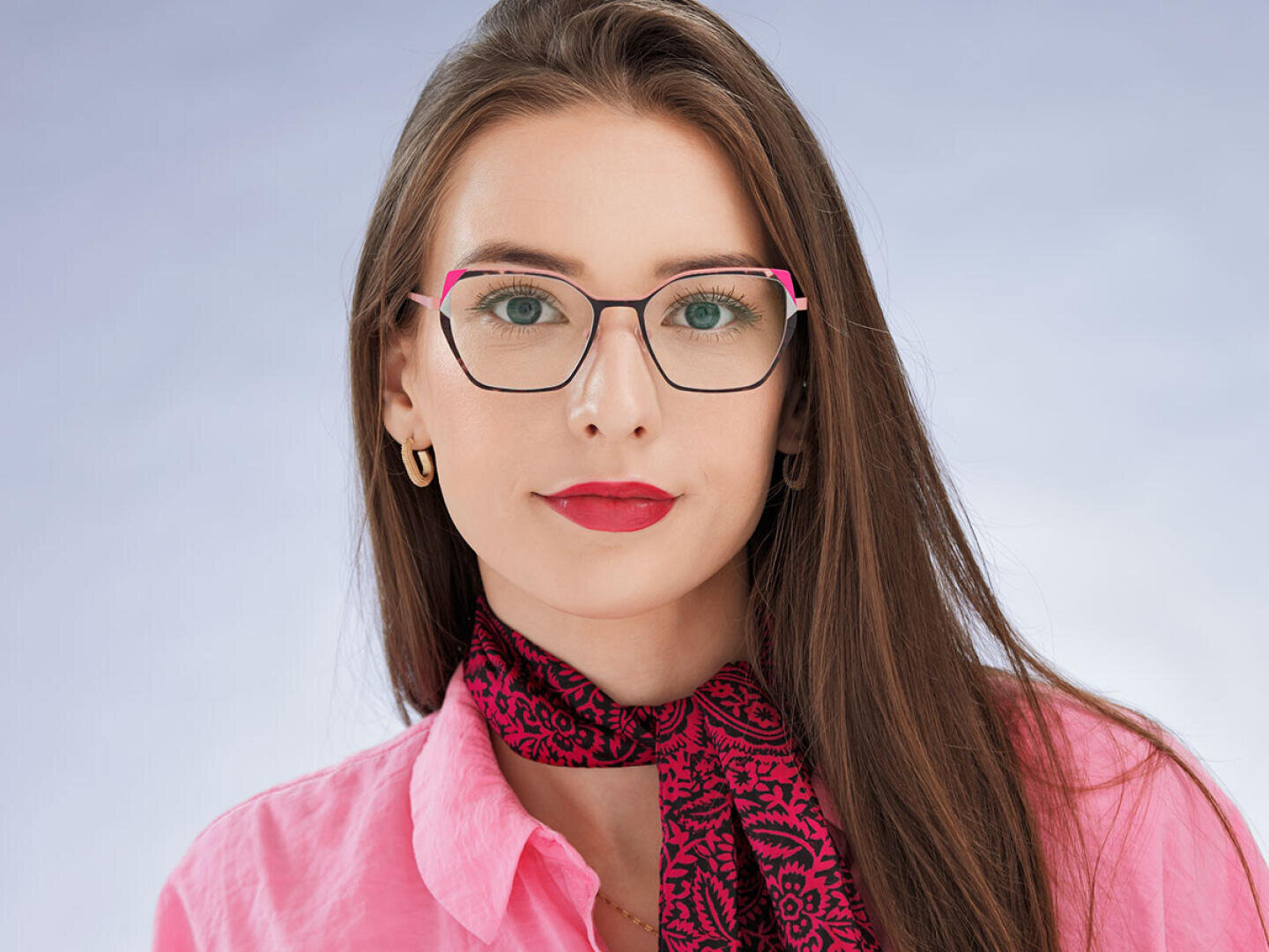 Eine Frau mit langen braunen Haaren, die eine Brille mit rosafarbenem Gestell, ein rosafarbenes Hemd und ein gemustertes rosa-schwarzes Halstuch trägt, posiert vor einem hellblauen Hintergrund. Sie trägt goldene Reifohrringe und roten Lippenstift.