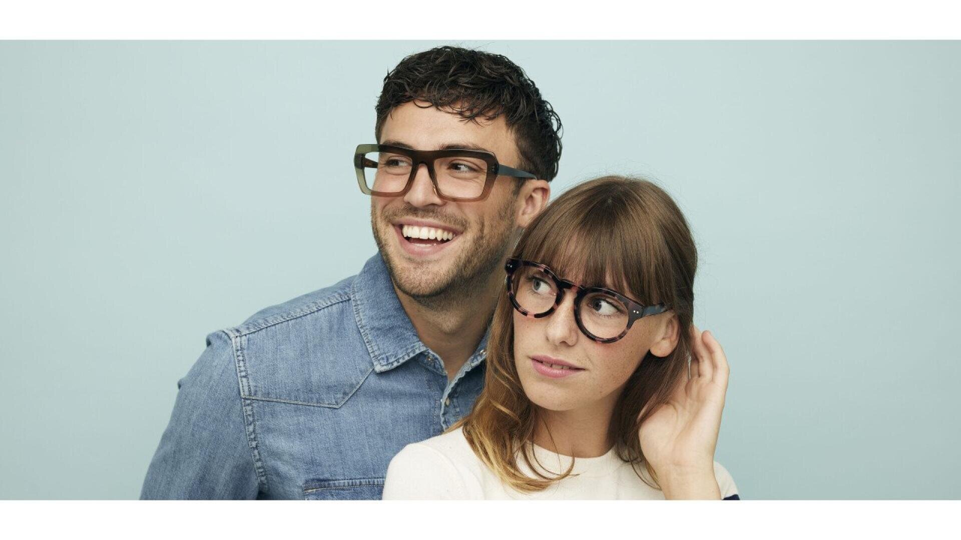 A man and a woman wearing large, stylish eyeglasses stand close together against a light blue background. The man smiles and looks to the side, while the woman looks up with a thoughtful expression, touching her hair.