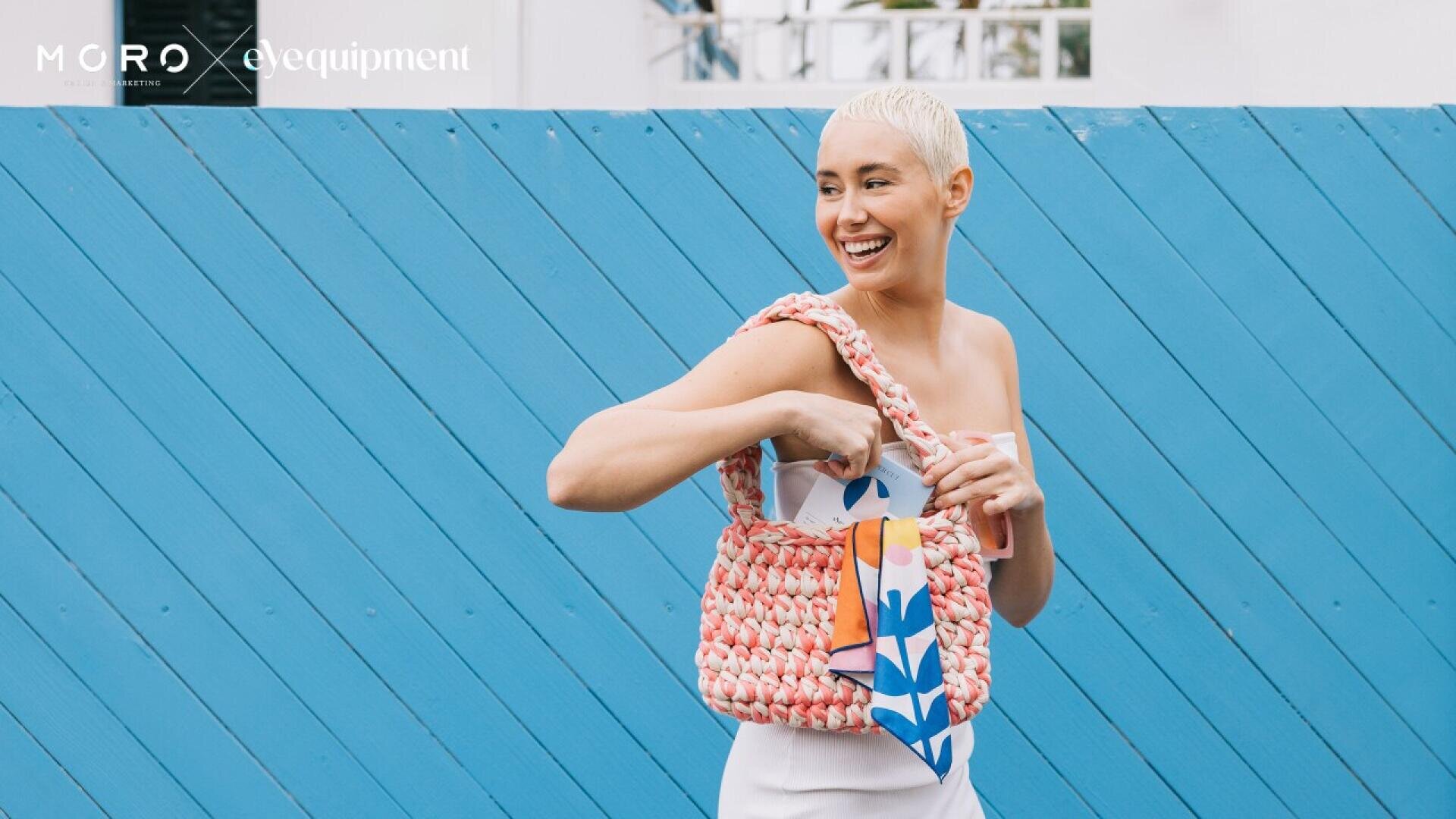 A smiling person with short blonde hair and a white outfit holds a pink woven handbag with a colorful scarf, standing in front of a bright blue wooden fence. Logos are visible in the top left corner.