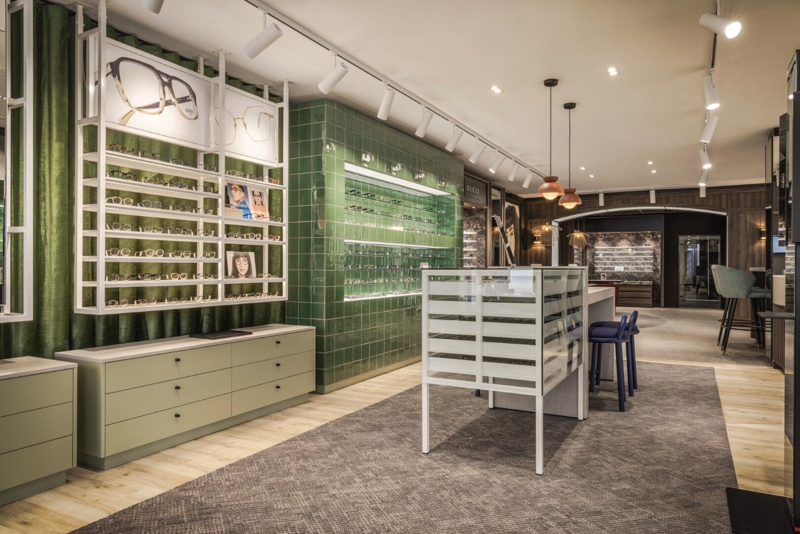 Modernes Brillengeschäft mit grün gekachelter Wand, Brillen in Regalen, einem zentralen Ausstellungstisch und stilvoller Beleuchtung. Der Raum hat helle Holzböden, grüne Akzente und eine komfortable, einladende Atmosphäre.