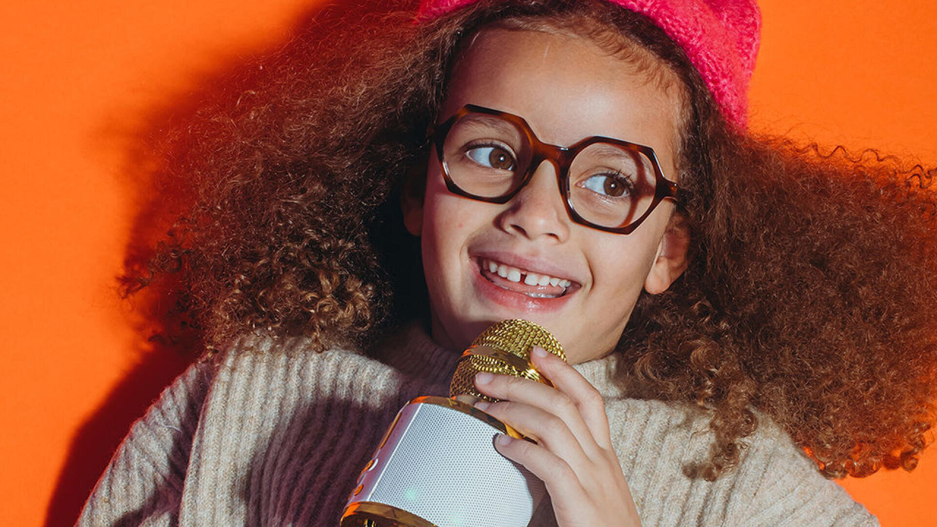 Ein lächelndes Kind mit lockigem Haar, Brille und rosa Mütze hält ein gold-weißes Mikrofon vor einem orangefarbenen Hintergrund.