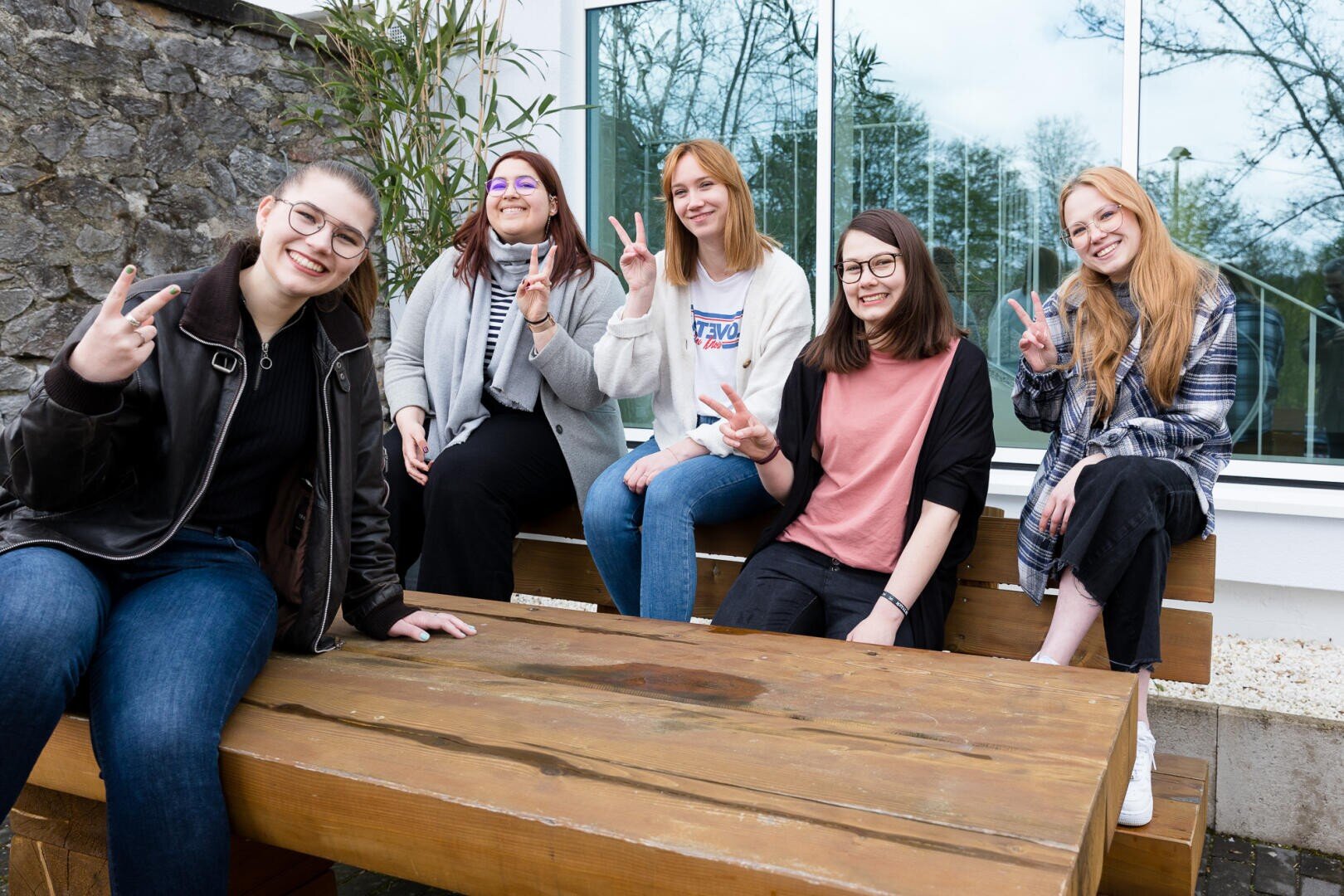 Fünf junge Frauen sitzen lächelnd auf Holzbänken im Freien und machen mit ihren Händen Friedenszeichen. Sie sind leger gekleidet und scheinen ihre gemeinsame Zeit in einer entspannten Umgebung zu genießen.