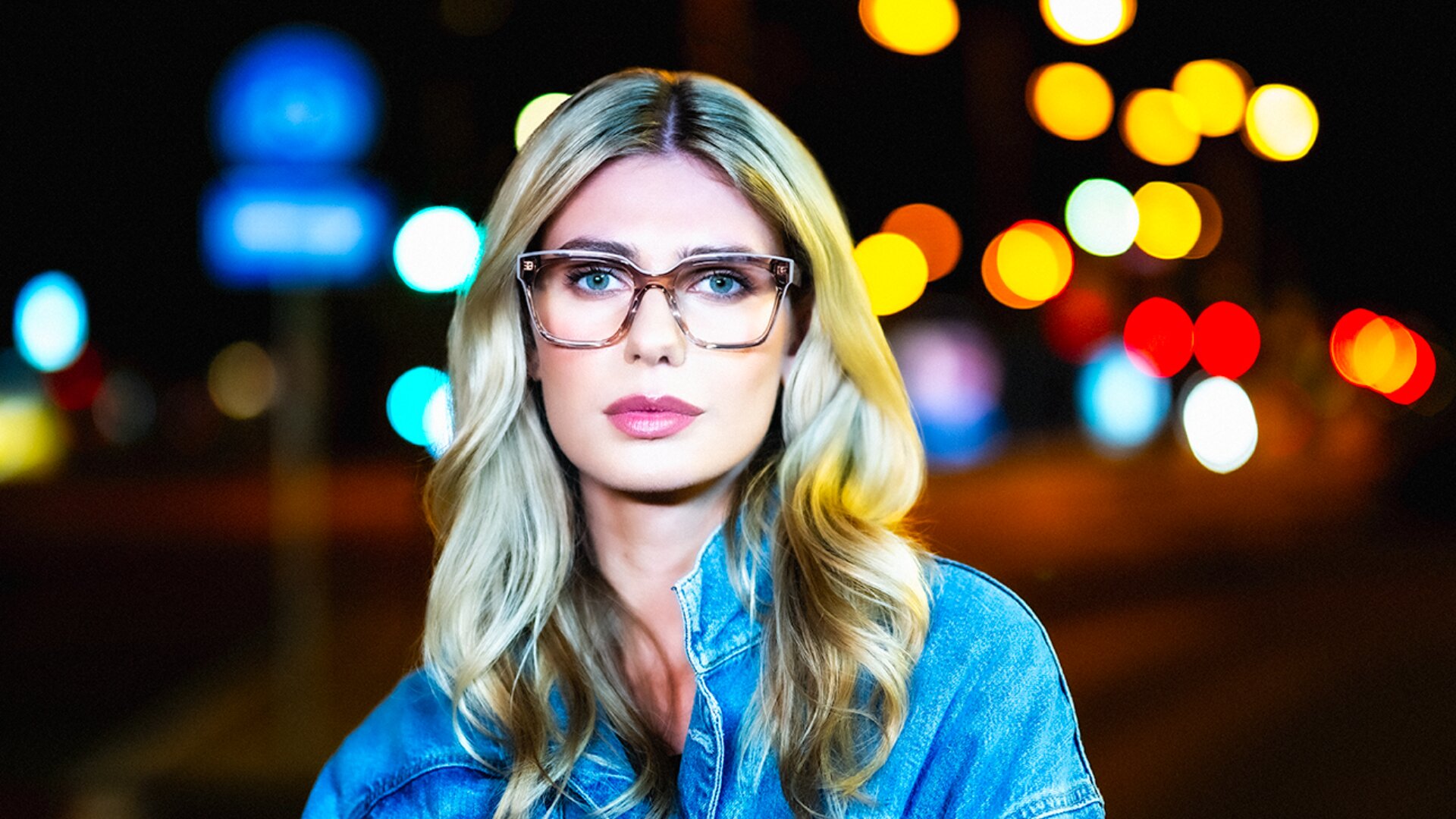 Eine Frau mit langen blonden Haaren und Brille trägt eine Jeansjacke und steht nachts im Freien, mit bunten, verschwommenen Lichtern der Stadt im Hintergrund.
