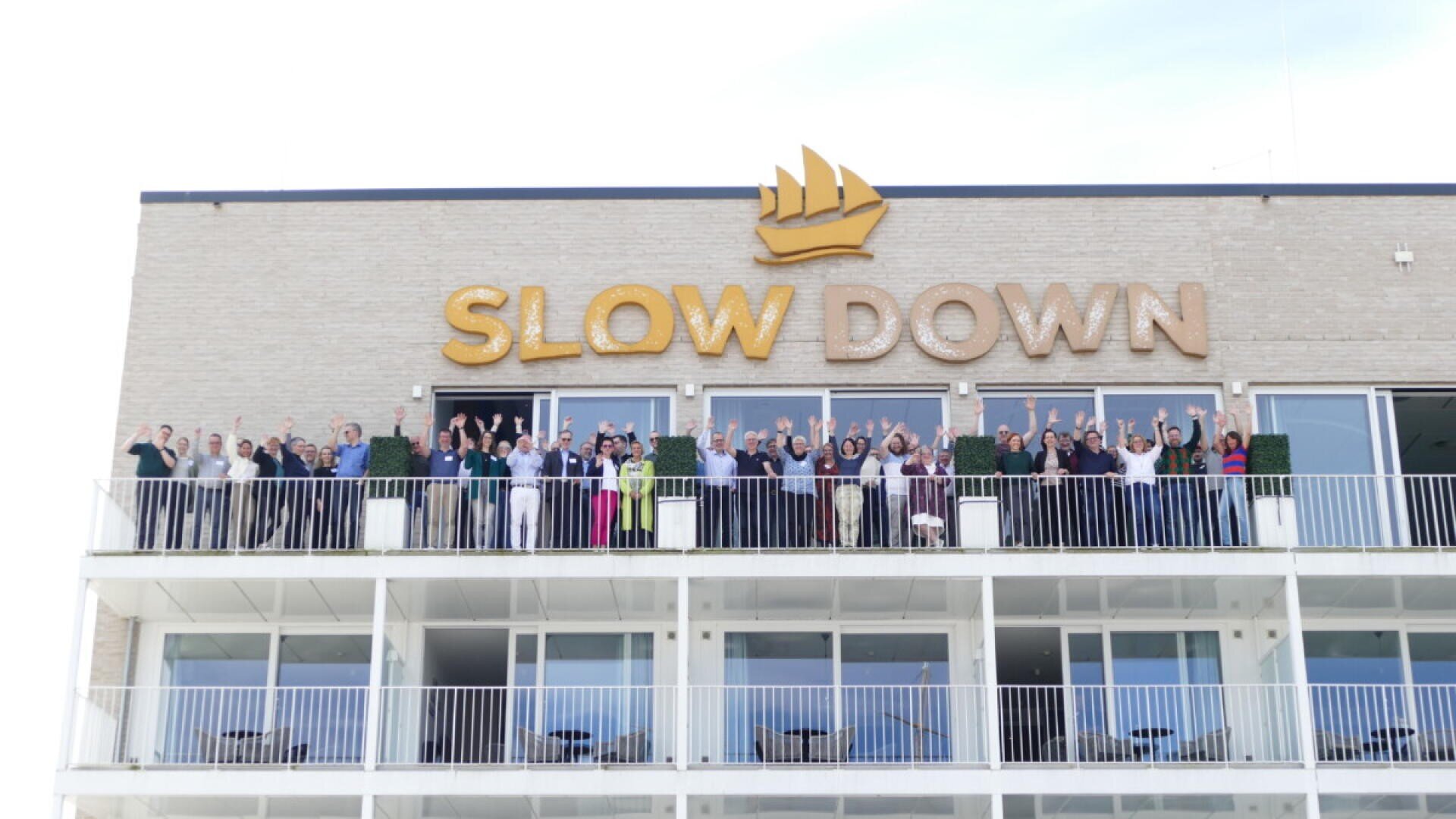 Eine Gruppe von Menschen steht lächelnd und mit erhobenen Armen auf einem Balkon vor einem Gebäude mit einem großen „SLOW DOWN“-Schild und einem goldenen Schiffslogo darüber.