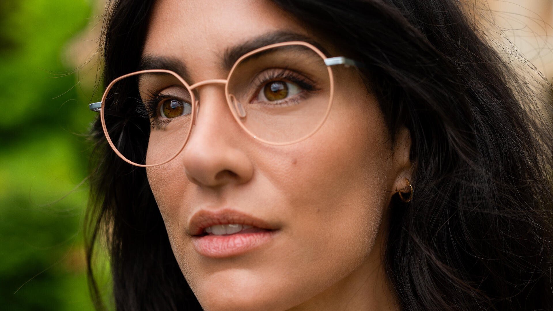 Nahaufnahme einer Frau mit langem, dunklem Haar und einer großen, runden Brille mit Goldrahmen. Sie hat hellbraune Augen, natürliches Make-up und trägt einen kleinen goldenen Creolen im linken Ohr. Grünes Laub ist im Hintergrund unscharf.