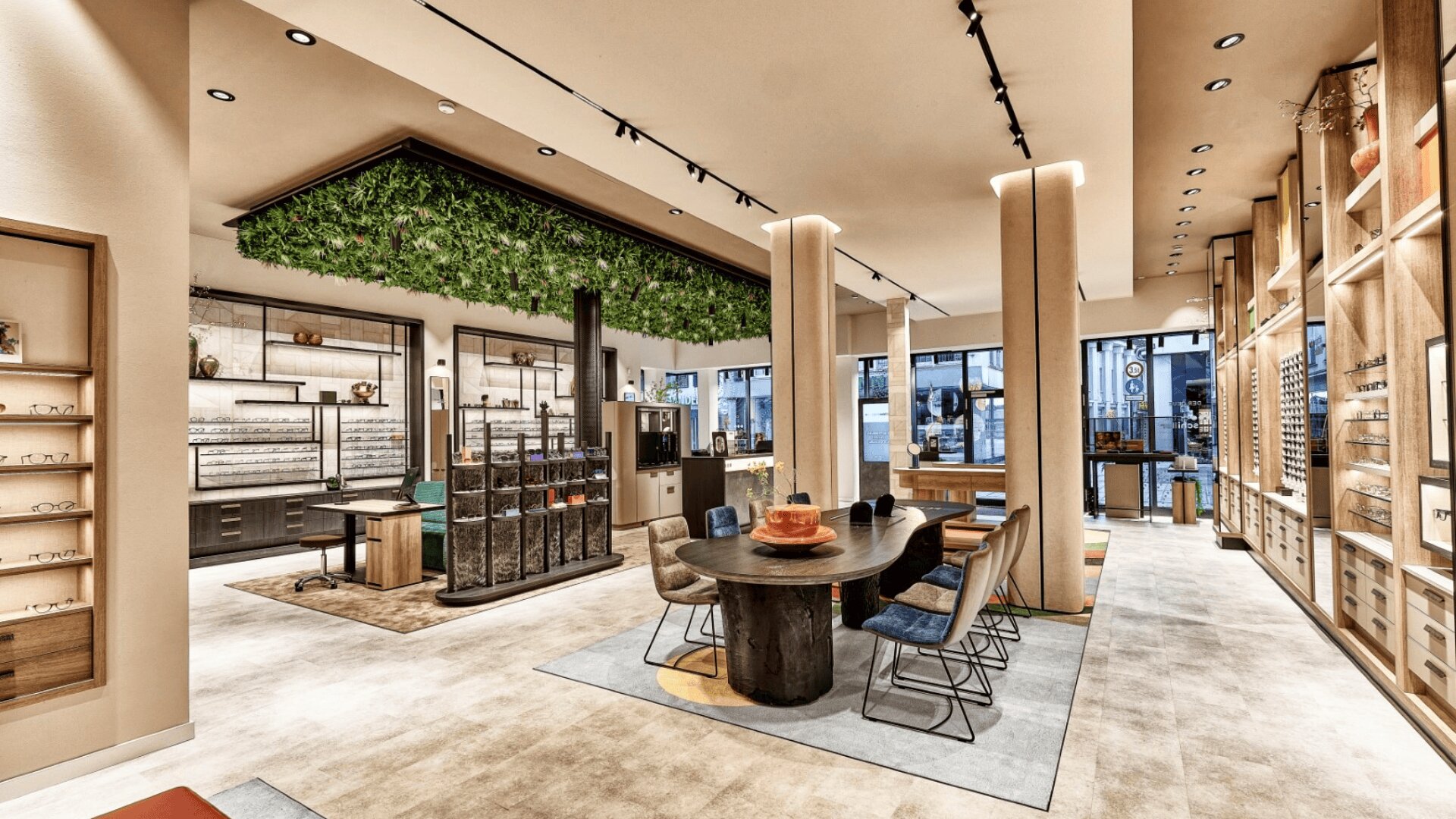 Modern eyewear store with stylish decor, including a central table with chairs, wall displays of eyeglasses, a ceiling feature with hanging greenery, and large windows letting in natural light.