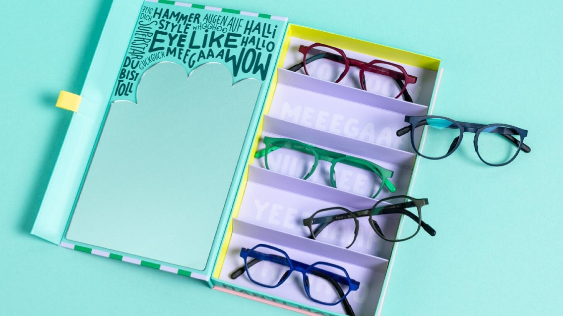 Eine pastellfarbene Schachtel mit fünf verschiedenfarbigen Brillen, die in separaten Fächern aufbewahrt werden, steht neben einem Notizblock mit verspielten Texten und Sätzen. Der Hintergrund ist in einem hellen Teeton gehalten.