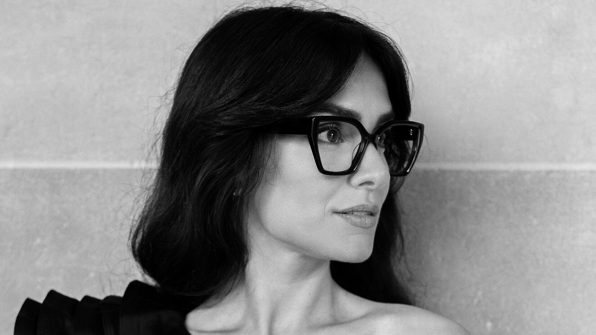 Schwarzweißfoto einer Frau mit langem, dunklem Haar, einer großen, eckigen Brille und einem schulterfreien Oberteil, die vor einer schlichten Wand nach rechts blickt.