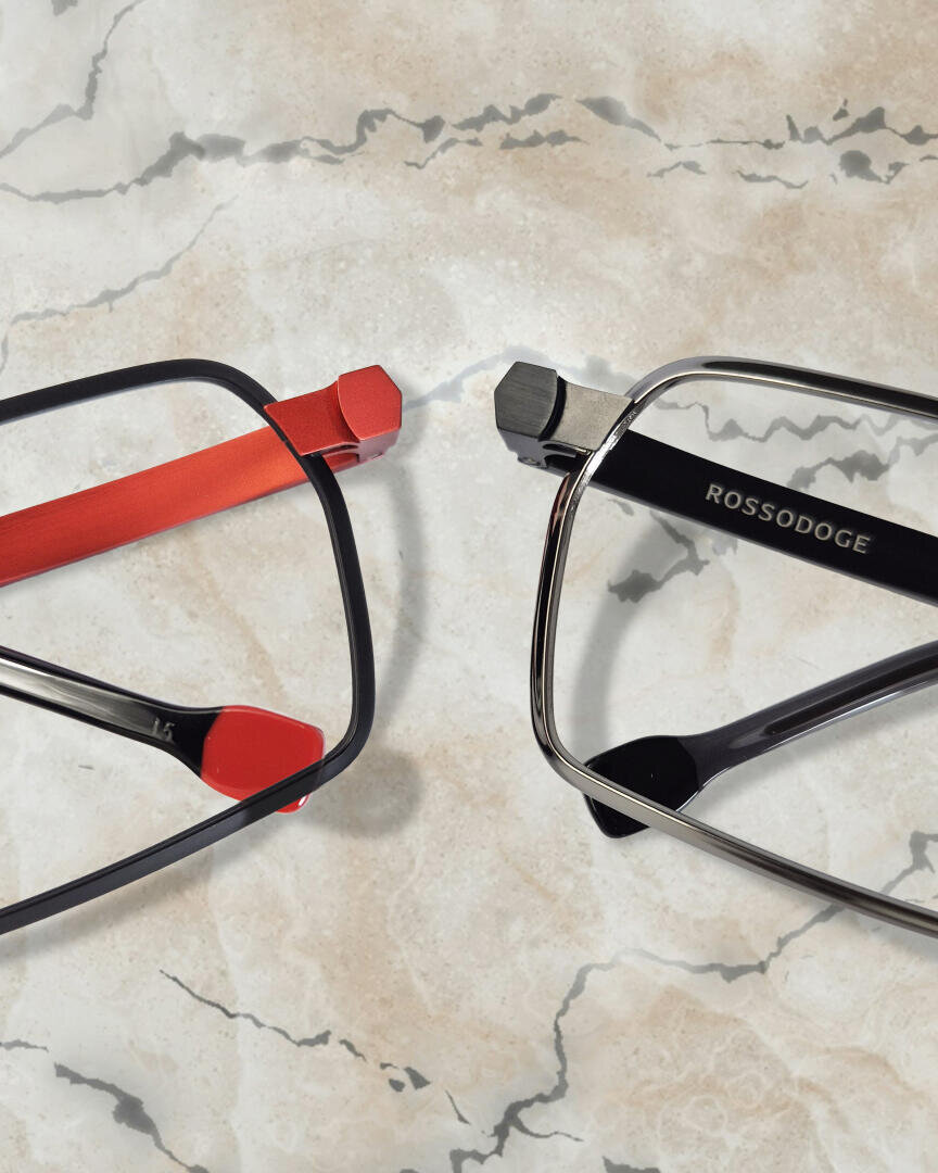 Two pairs of rectangular eyeglasses on a marble surface: one with red and black frames, the other with black and silver frames labeled ROSSODOGE.