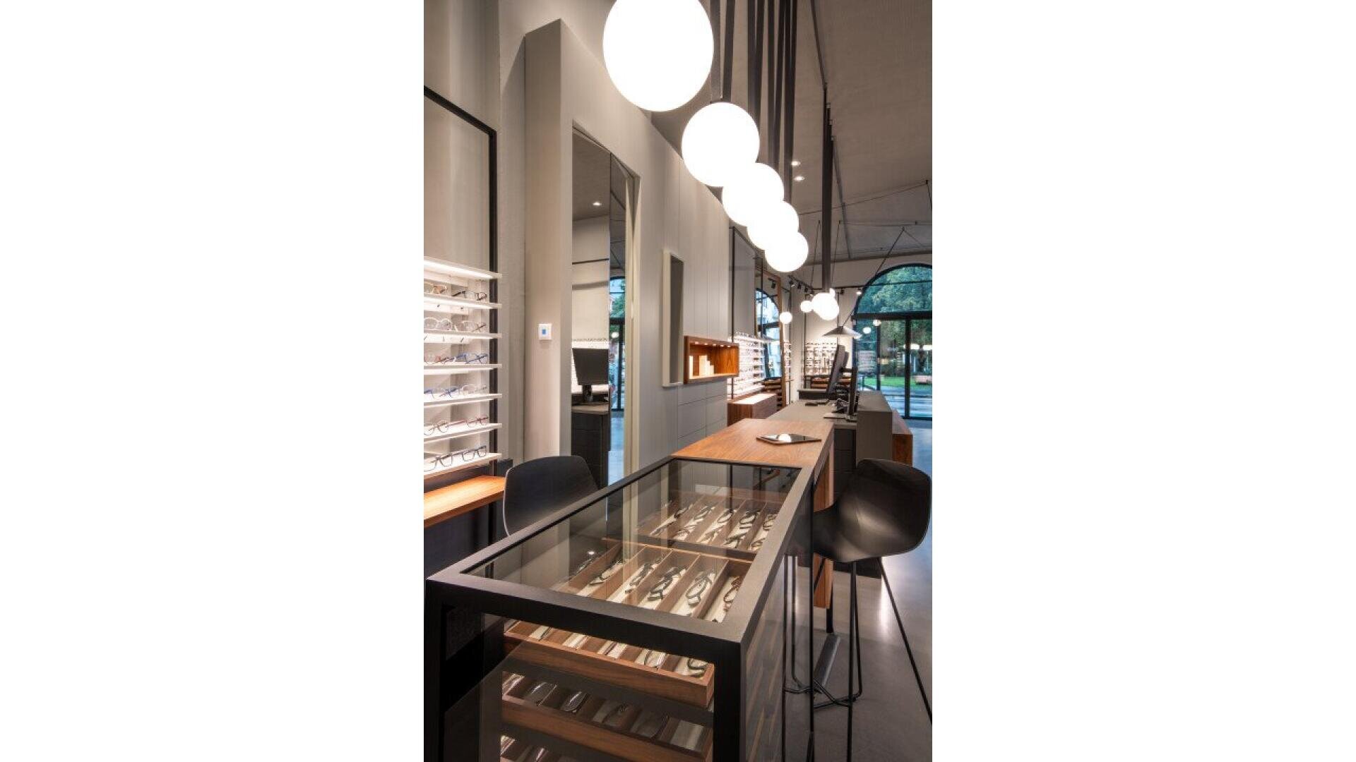 Modern eyewear store interior with a long glass display case of eyeglasses, black bar stools, spherical pendant lights hanging above, wall mirrors, and large windows letting in natural light.