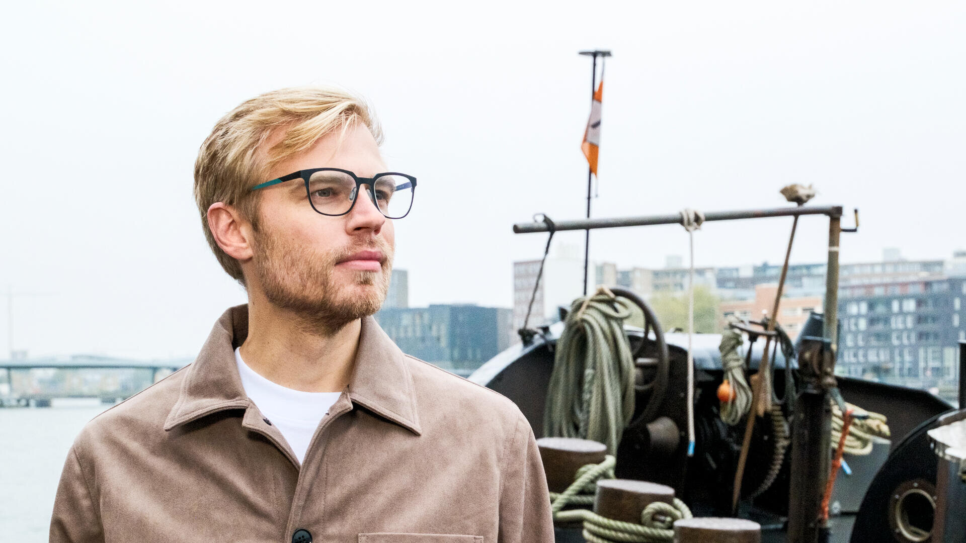 A man with blond hair, a beard, and glasses stands outdoors near a docked boat, looking into the distance. Buildings and water are visible in the background on an overcast day.