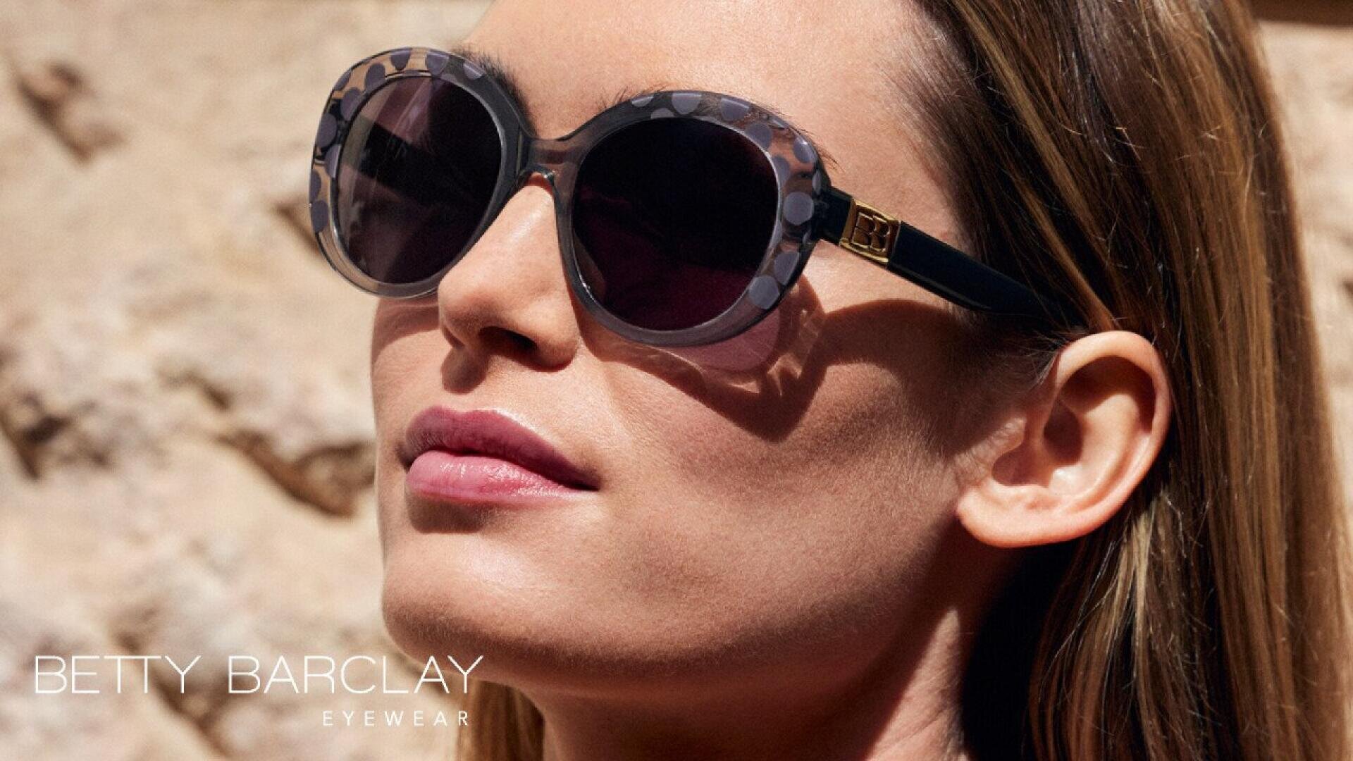 A woman wearing large, round sunglasses with patterned frames looks upward. The background is sunlit stone. Betty Barclay Eyewear text appears in the bottom left corner.