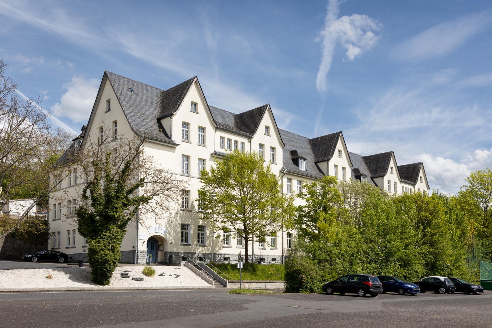 Ein großes, weißes, mehrstöckiges Gebäude mit steilen, dunkelgrauen Dächern und Giebeln steht hinter belaubten Bäumen. Davor sind mehrere Autos entlang der Straße geparkt, und der Himmel ist blau mit einigen Wolken.