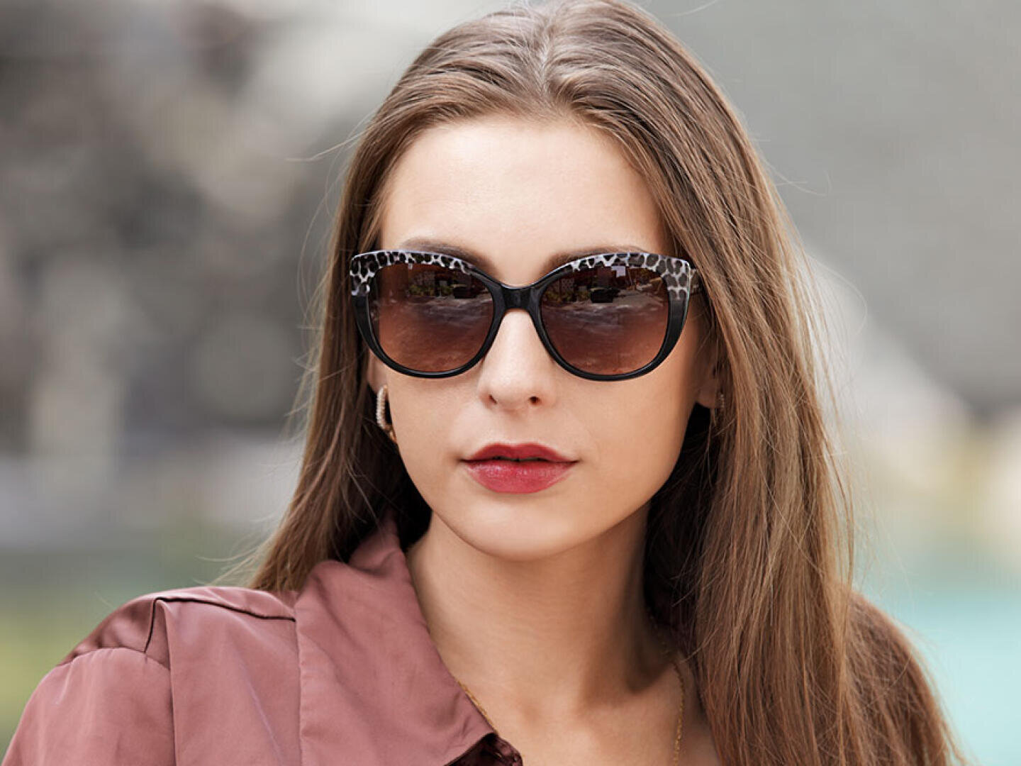 Eine Frau mit langen braunen Haaren, die eine große, gemusterte Sonnenbrille und eine lilafarbene Bluse trägt, blickt leicht zur Seite und hat einen neutralen Ausdruck. Der Hintergrund ist unscharf.