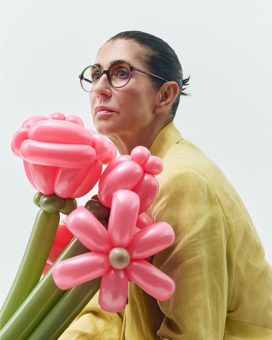 A woman with glasses and dark hair tied back, wearing a light yellow jacket, sits next to large pink balloon flowers with green stems against a plain light background.