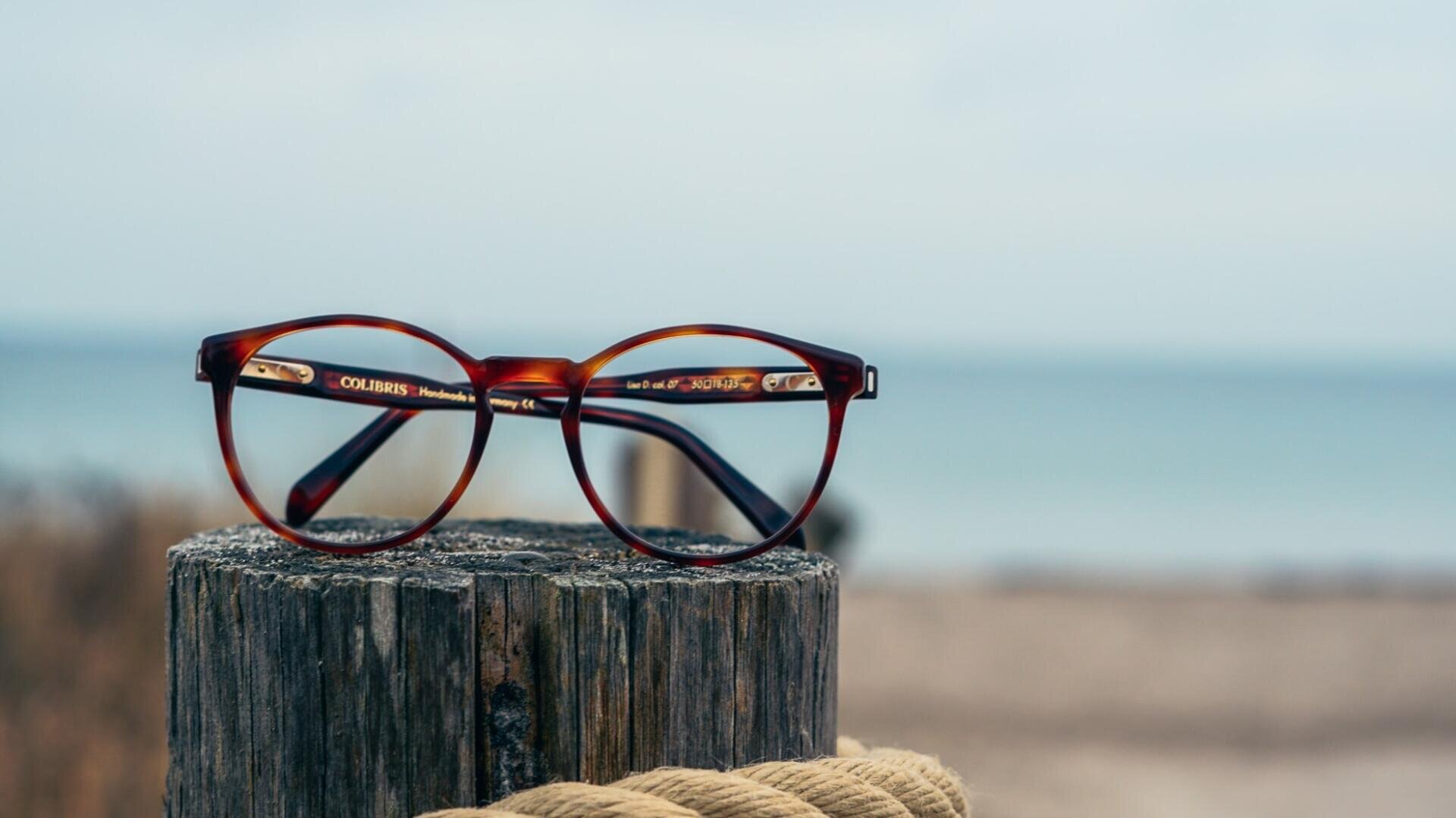 Eine braune, runde Brille ruht auf einem verwitterten Holzpfosten mit einem verschwommenen Strand und Meer im Hintergrund.