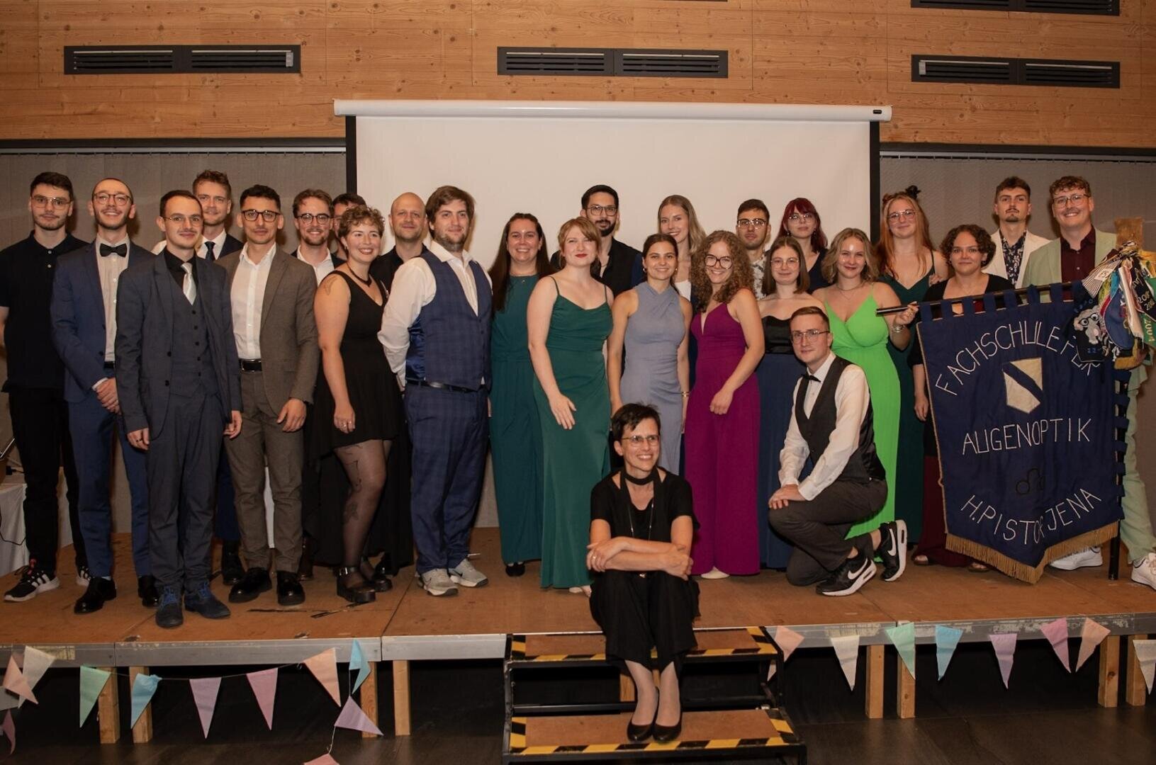 Eine Gruppe von formell gekleideten Personen posiert gemeinsam auf einer Bühne vor einem Bildschirm. Eine Frau sitzt vorne am Rand der Bühne, während andere hinter ihr stehen. Auf einem Transparent steht Fachschule für Augenoptik Jena.