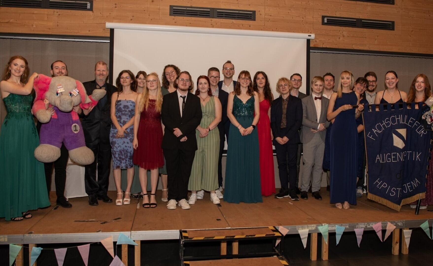 Eine Gruppe förmlich gekleideter Menschen steht auf einer Bühne vor einem Bildschirm. Einige halten einen großen ausgestopften Elefanten und ein geschmücktes Transparent. Der Hintergrund ist aus Holz, und alle lächeln für das Foto.