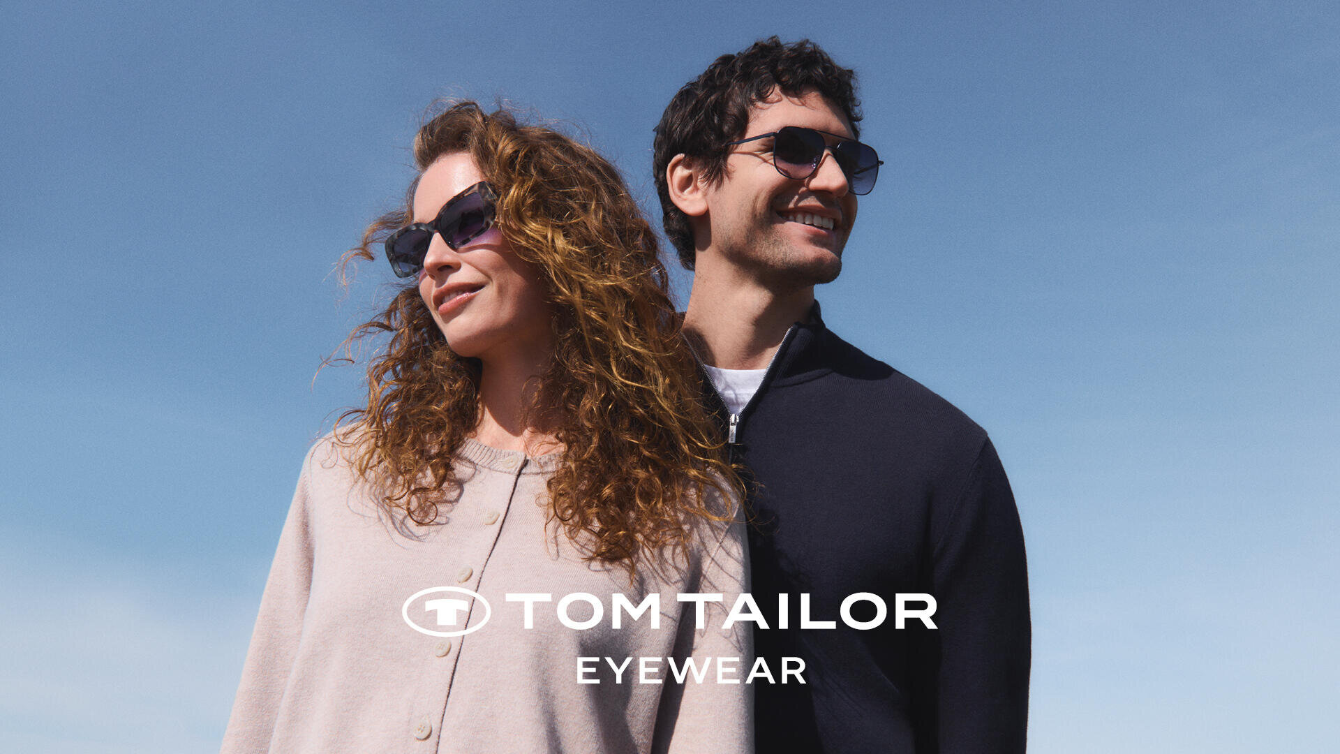 Eine Frau und ein Mann mit Sonnenbrille stehen Rücken an Rücken und lächeln vor einem blauen Himmel. Das Tom Tailor Eyewear-Logo ist am unteren Rand des Bildes zu sehen.