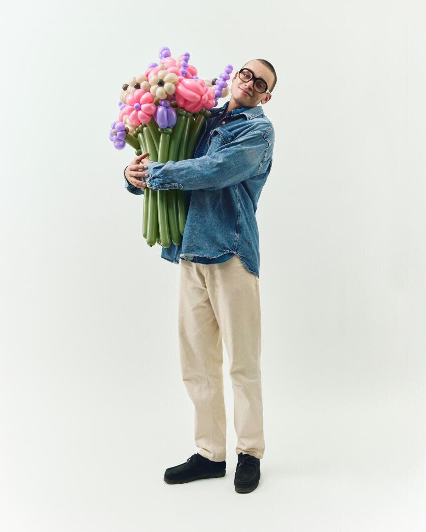 A smiling person wearing glasses, a denim jacket, beige trousers and black shoes holds a large, colorful bouquet of twisted balloons in front of a plain white background.