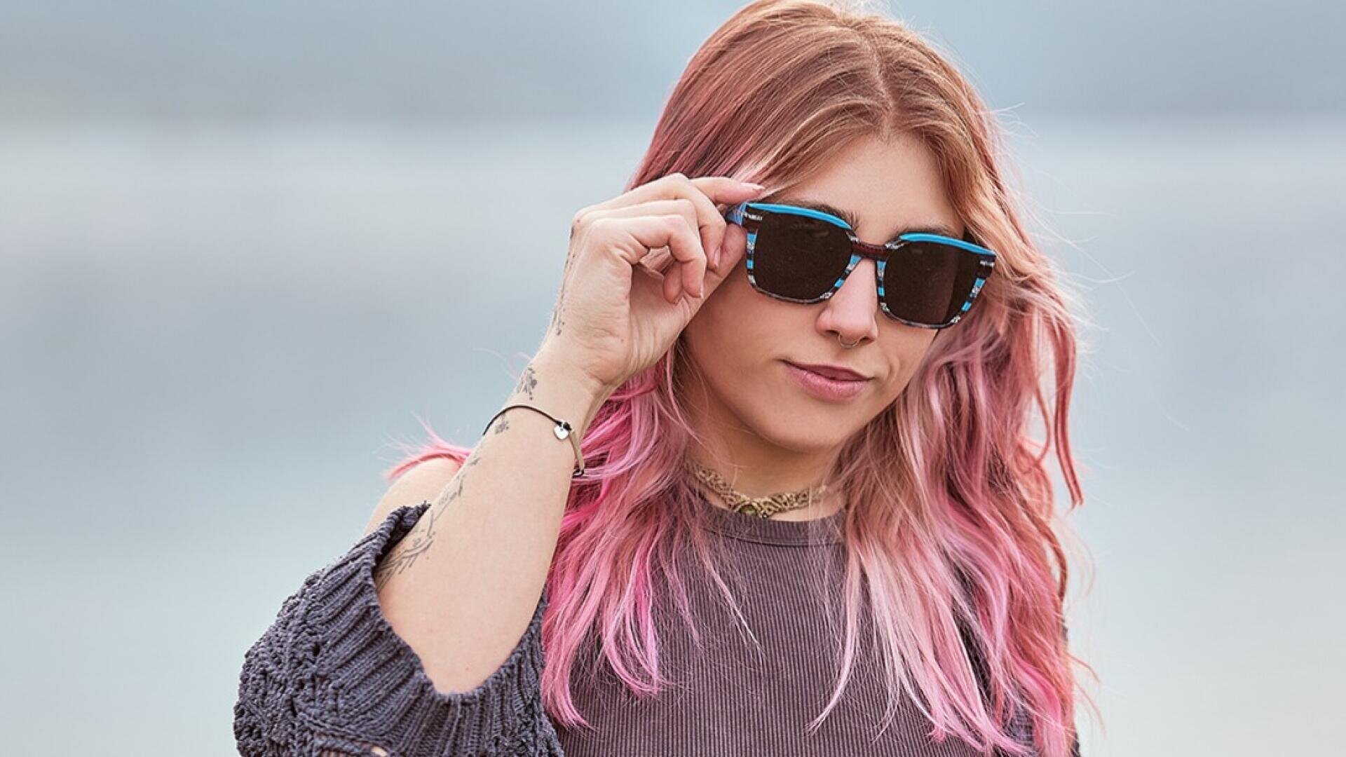A person with long, wavy pink hair, wearing black sunglasses and a gray sweater, stands outdoors, holding sunglasses in one hand, with a calm, blurred background in the background.