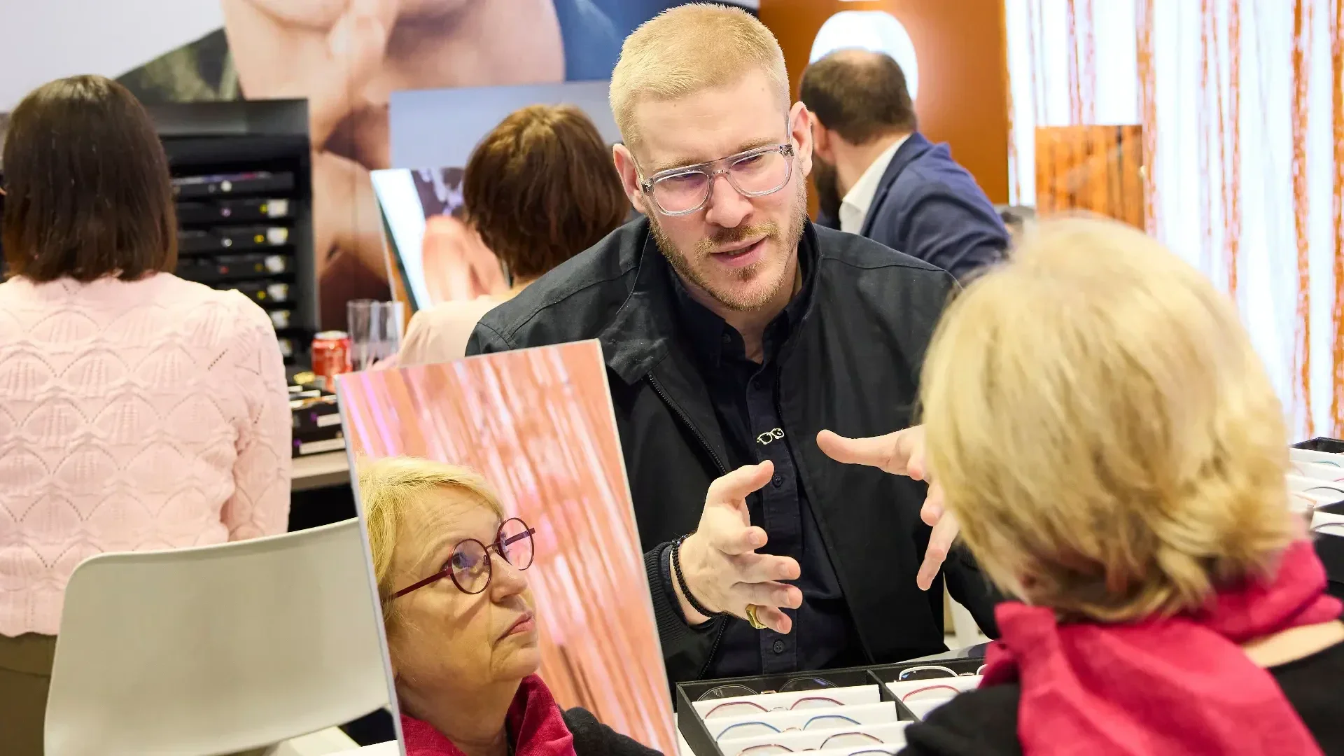 A man with glasses gestures while speaking to an older woman, whose reflection is visible in a mirror on the table. They are surrounded by eyeglasses, suggesting an eyewear fitting or consultation in a busy store.