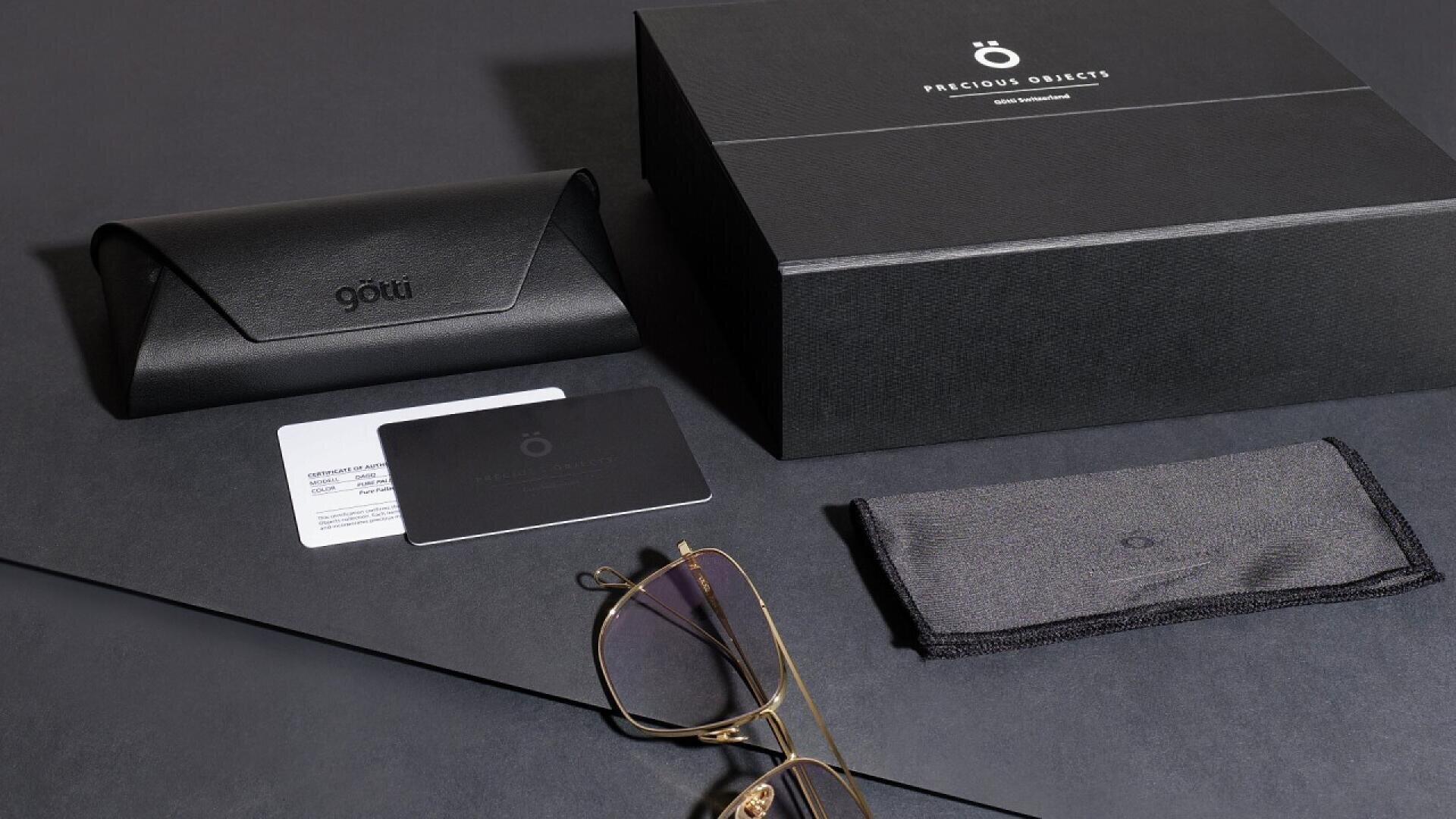 A pair of gold-framed eyeglasses, a black leather case, a black cleaning cloth, two cards, and a large black box labeled Precious Objects are arranged on a dark surface.