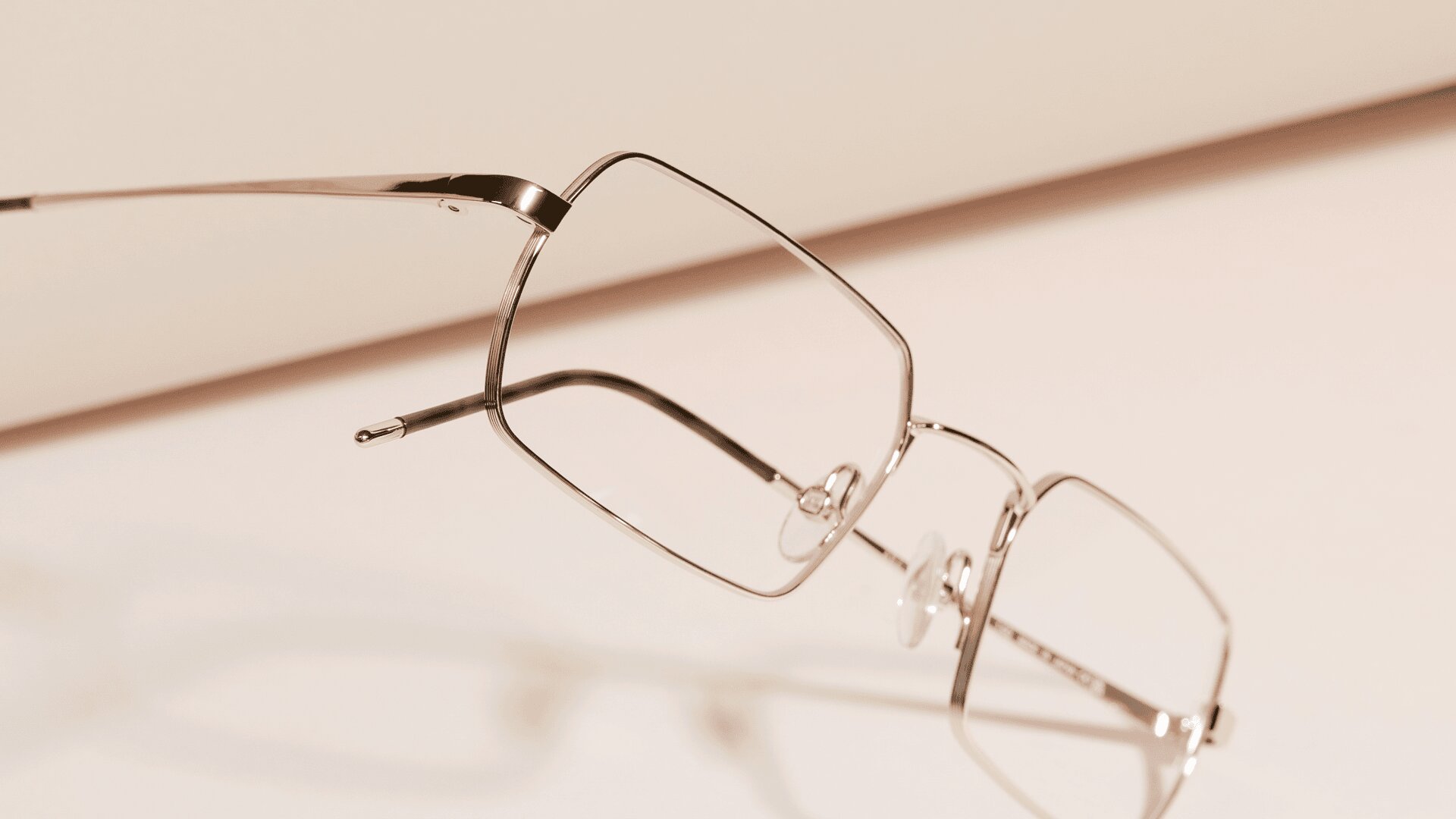 Eine Nahaufnahme einer Metallbrille mit rechteckigem Rahmen, die auf einer hellen, cremefarbenen Oberfläche liegt und einen weichen Schatten wirft. Das Bild hat eine minimalistische und elegante Ausstrahlung.