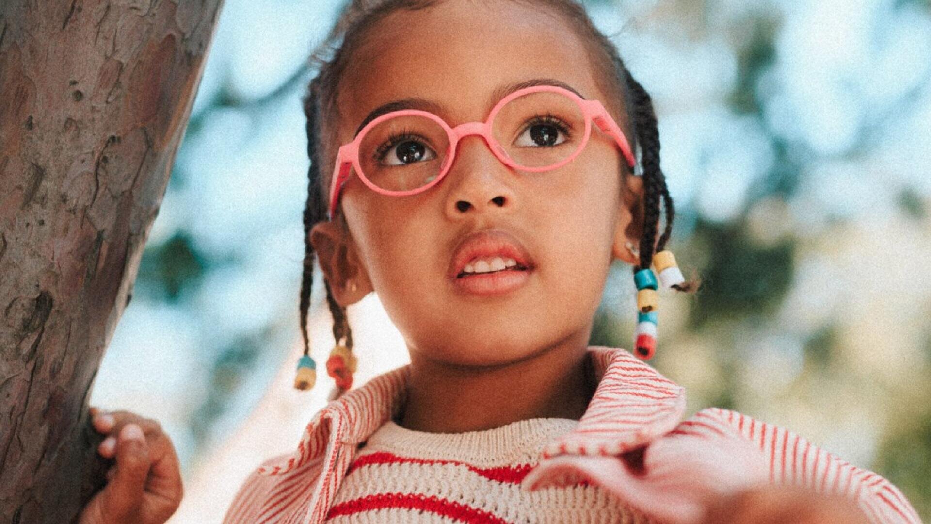 A young child with braids and colorful beads wears pink glasses and a red-and-white striped shirt, looking slightly upward while standing outdoors near a tree, with sunlight and blurred greenery in the background.