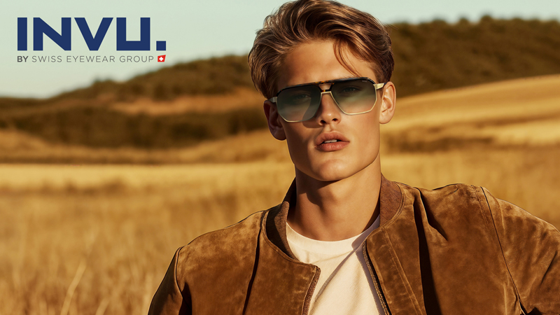 Ein junger Mann mit modischer Sonnenbrille und einer braunen Wildlederjacke steht auf einem sonnenbeschienenen Feld mit Hügeln im Hintergrund. Das Logo der INVU. BY SWISS EYEWEAR GROUP ist in der oberen linken Ecke zu sehen.