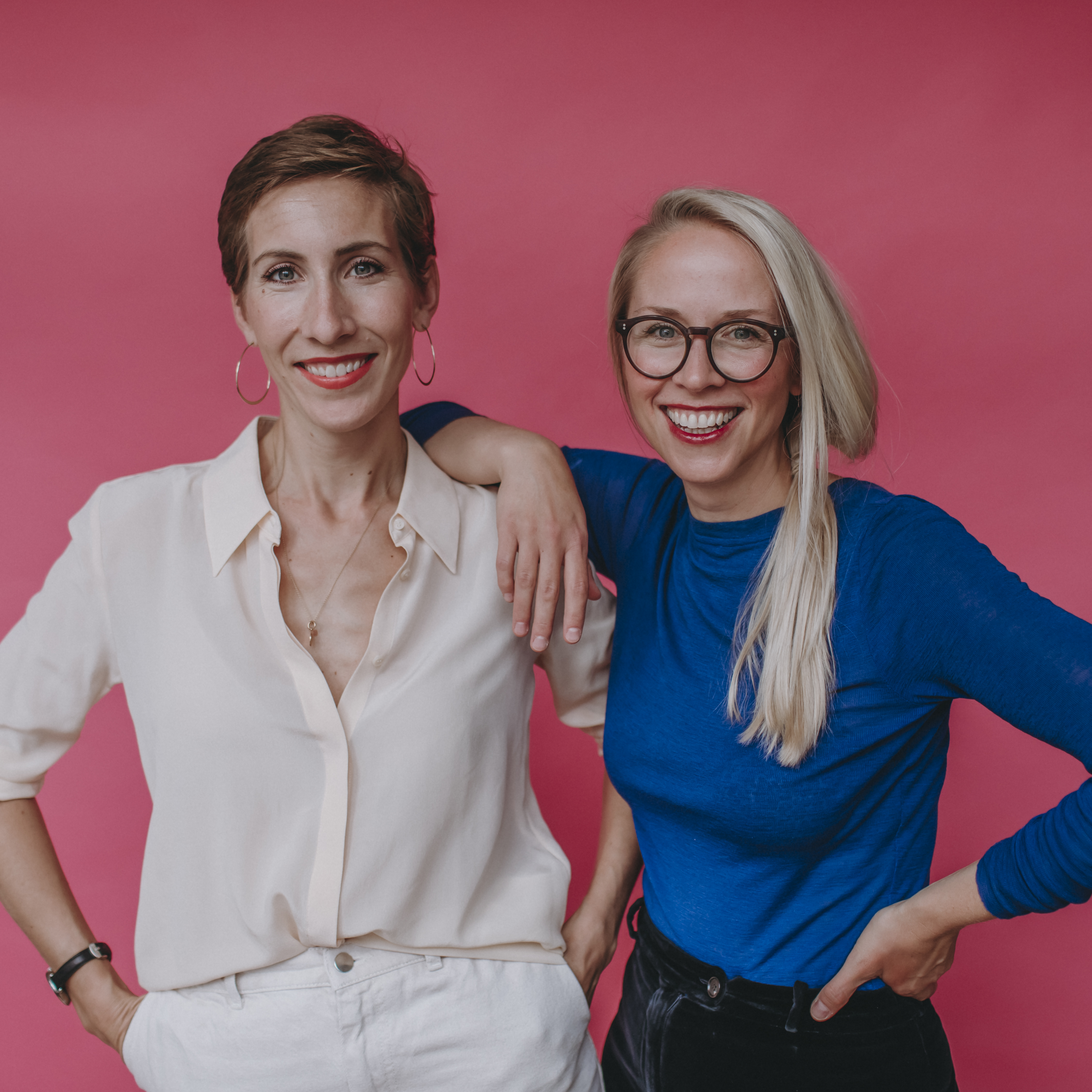 Zwei Frauen stehen vor einem rosa Hintergrund und lächeln. Die eine hat kurzes braunes Haar, Ohrringe, eine weiße Bluse und eine weiße Hose. Die andere hat lange blonde Haare, eine Brille, ein blaues, glitzerndes Oberteil und eine schwarze Hose.