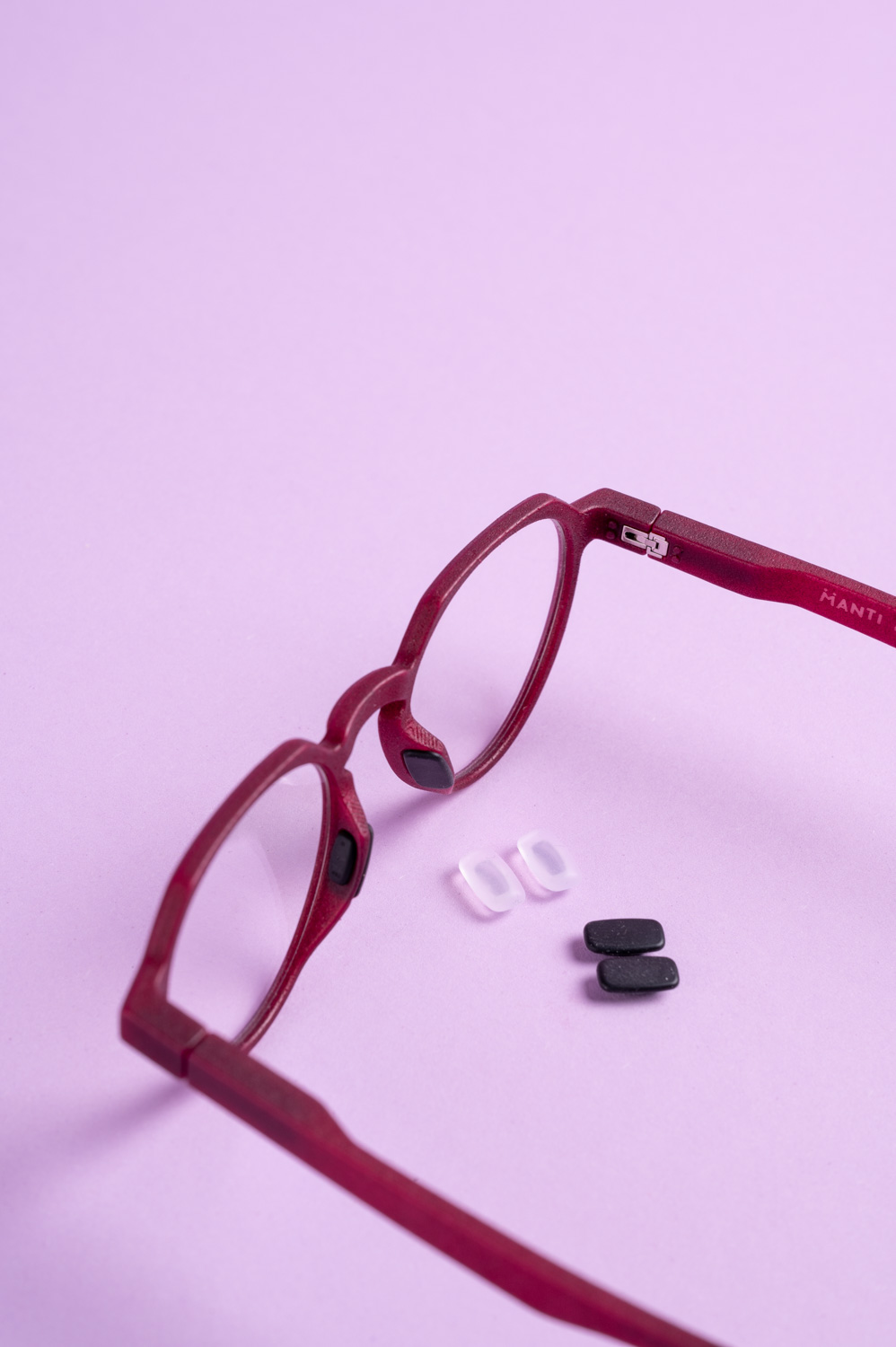 Red eyeglasses with black nose pads placed near two pairs of replacement nose pads, one black and one clear, on a light purple surface.