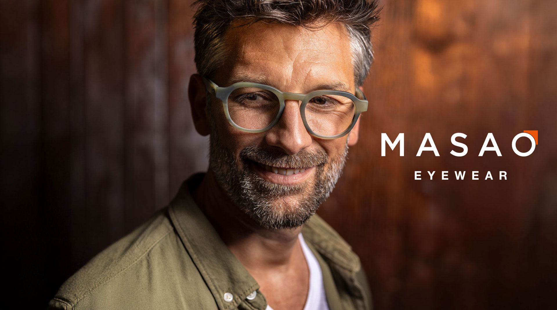 Ein l&auml;chelnder Mann mit grauem Haar und Bart tr&auml;gt eine hellgr&uuml;ne Brille und ein hellbraunes Hemd und steht vor einem dunklen Holzhintergrund. Neben ihm erscheint der Text MASAO EYEWEAR.