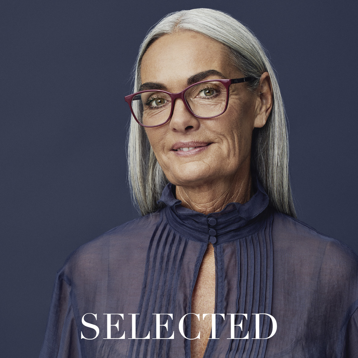 Eine ältere Frau mit langen grauen Haaren, einer roten Brille und einer hochgeschlossenen, marineblauen Bluse steht vor einem dunklen Hintergrund. Sie lächelt sanft. Das Wort SELECTED erscheint am unteren Rand des Bildes.
