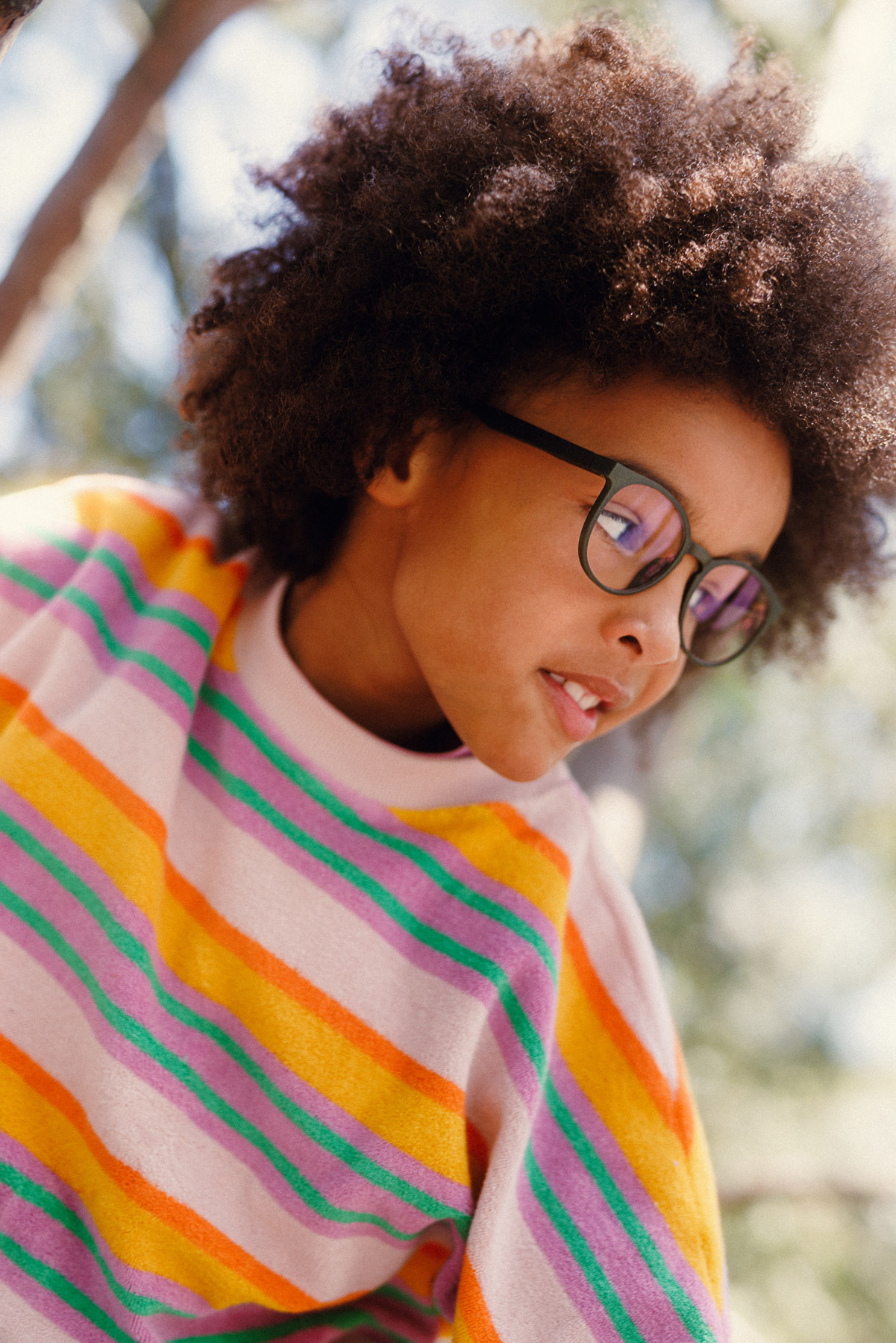 A child with curly hair and glasses wearing a colorful striped sweater looks downward, smiling softly. The background is outdoors with blurred trees and sunlight.