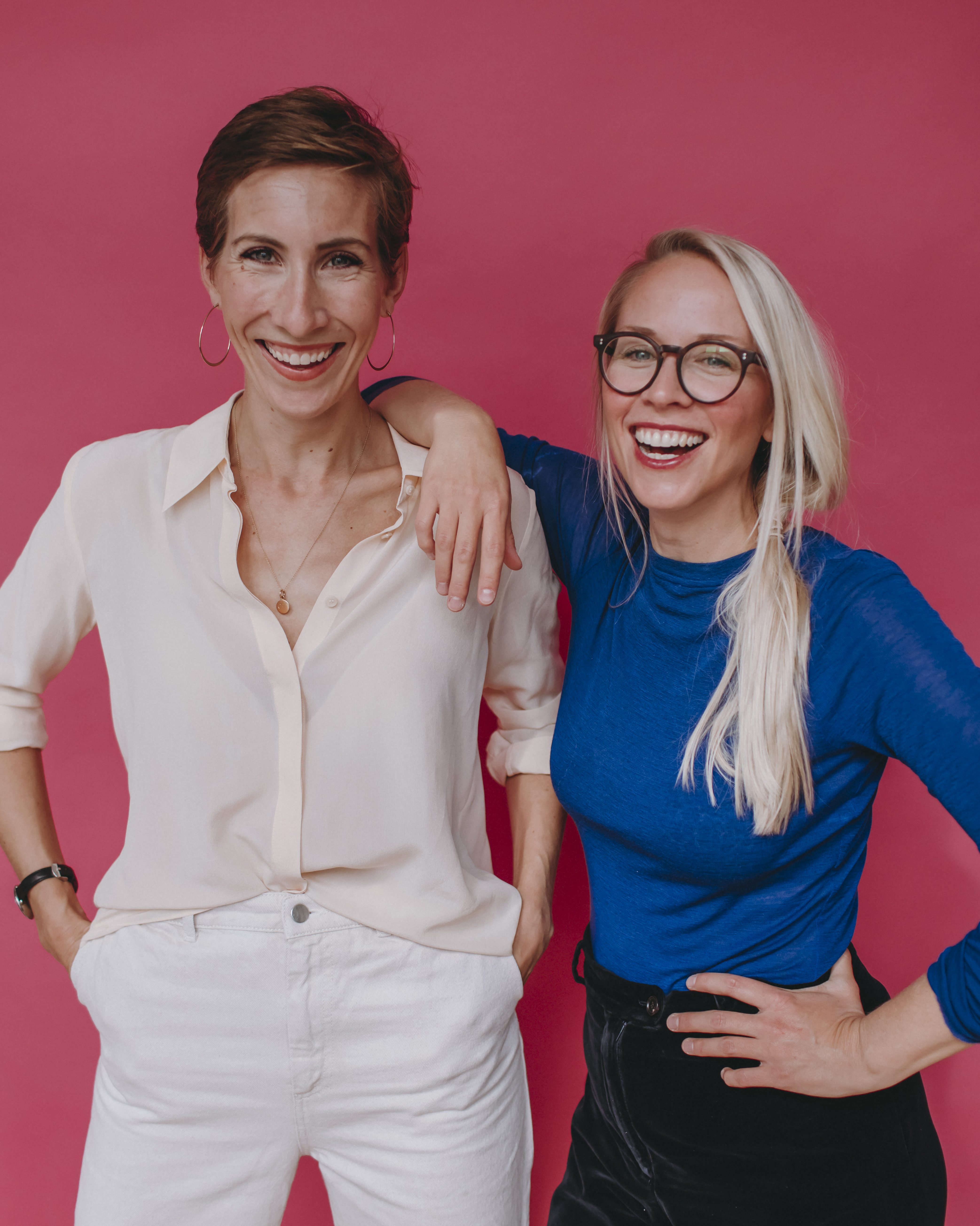 Two women smiling in front of a pink background. One has short brown hair, wears a light blouse and white pants; the other has long blond hair, glasses, and a blue top. They stand close, looking cheerful and relaxed.