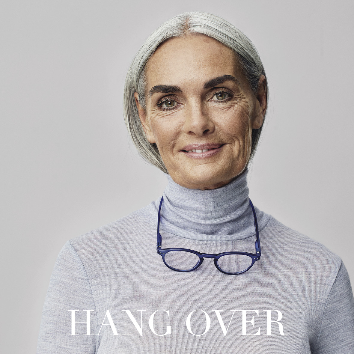Lächelnde ältere Frau mit grauem Haar und hellgrauem Rollkragenpullover, blaue Brille um den Hals und die Worte HANG OVER in fetter weißer Schrift am unteren Rand.