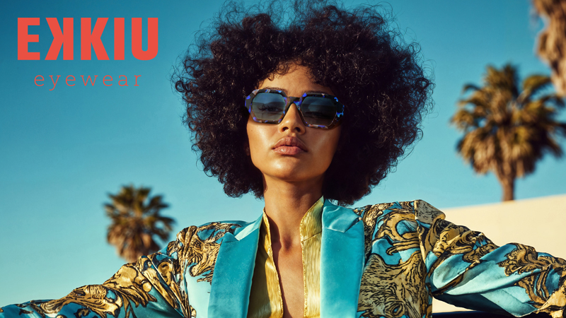 Eine stilvolle Frau mit lockigem Haar trägt eine große Sonnenbrille und eine gewagte, bunte Jacke im Freien, mit Palmen und blauem Himmel im Hintergrund. Der Text EKKIU eyewear erscheint in der oberen linken Ecke.