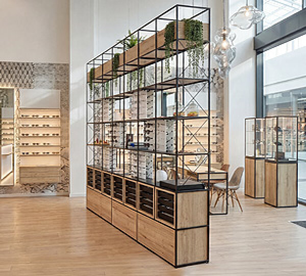 Modernes Brillengeschäft mit Holzböden, offenen Brillenregalen, Hängepflanzen und großen Fenstern, die natürliches Licht hereinlassen. Der Raum hat ein sauberes, minimalistisches und organisiertes Design.